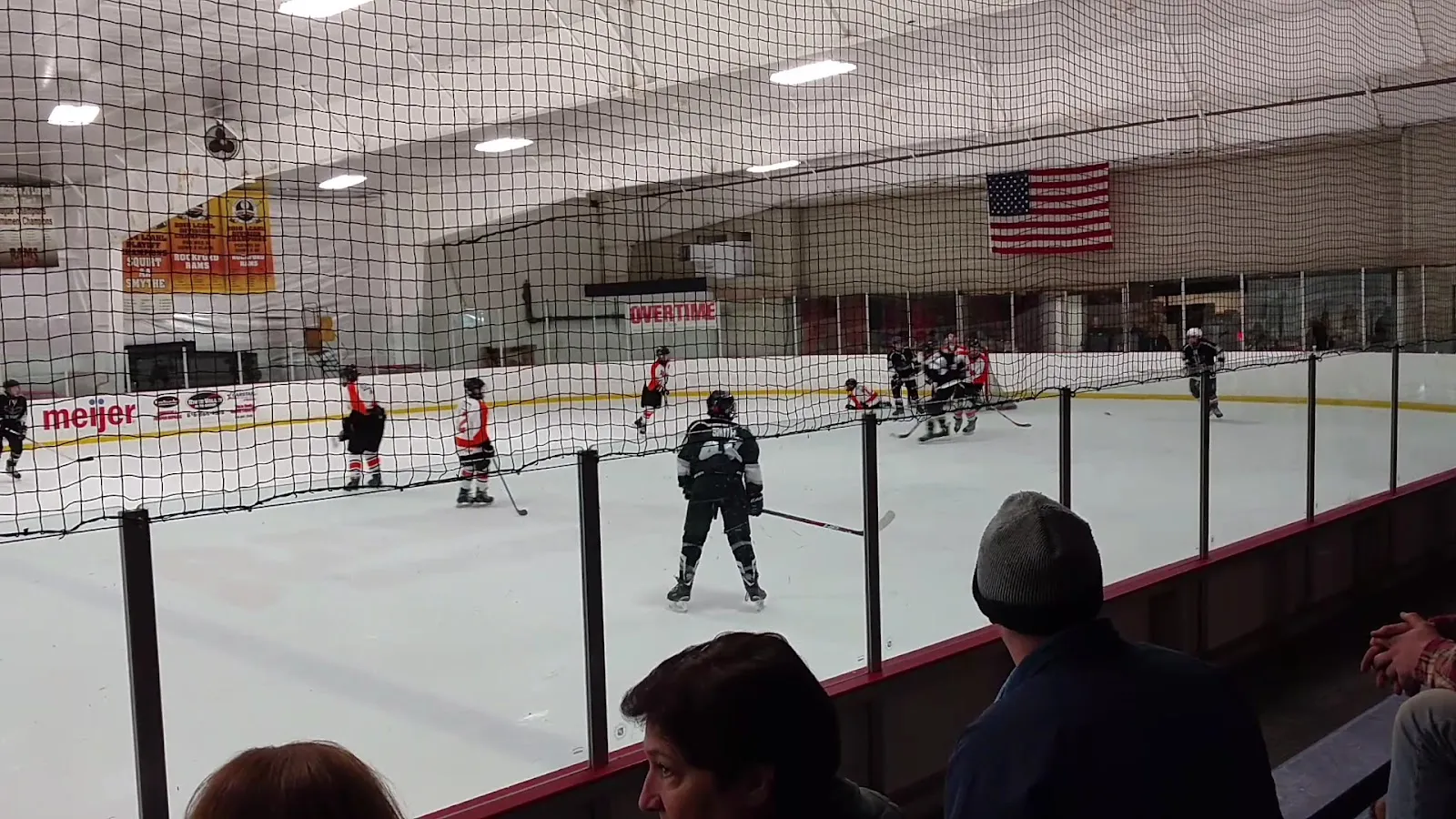 A youth game seen from the stands, players spread along the far boards under a high white roof with an American flag and sponsor boards on the end wall, spectators' heads at the bottom corners. — Cedar Rock Sports Plex