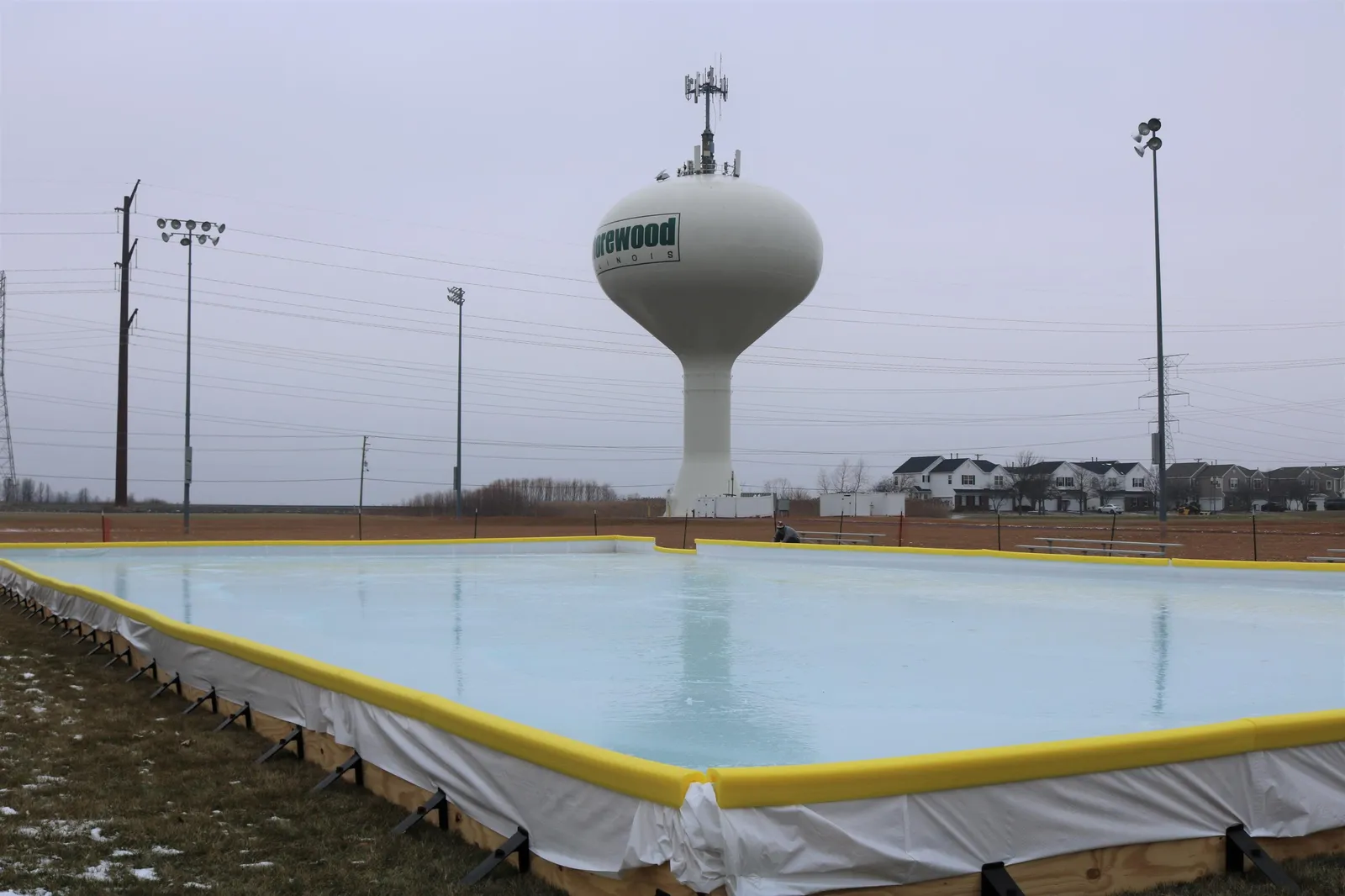 The outdoor ice rink at Cenes Four Seasons Park in Shorewood, with a yellow-edged liner and the village water tower behind it. — Cene's Four Seasons Park Ice Rink