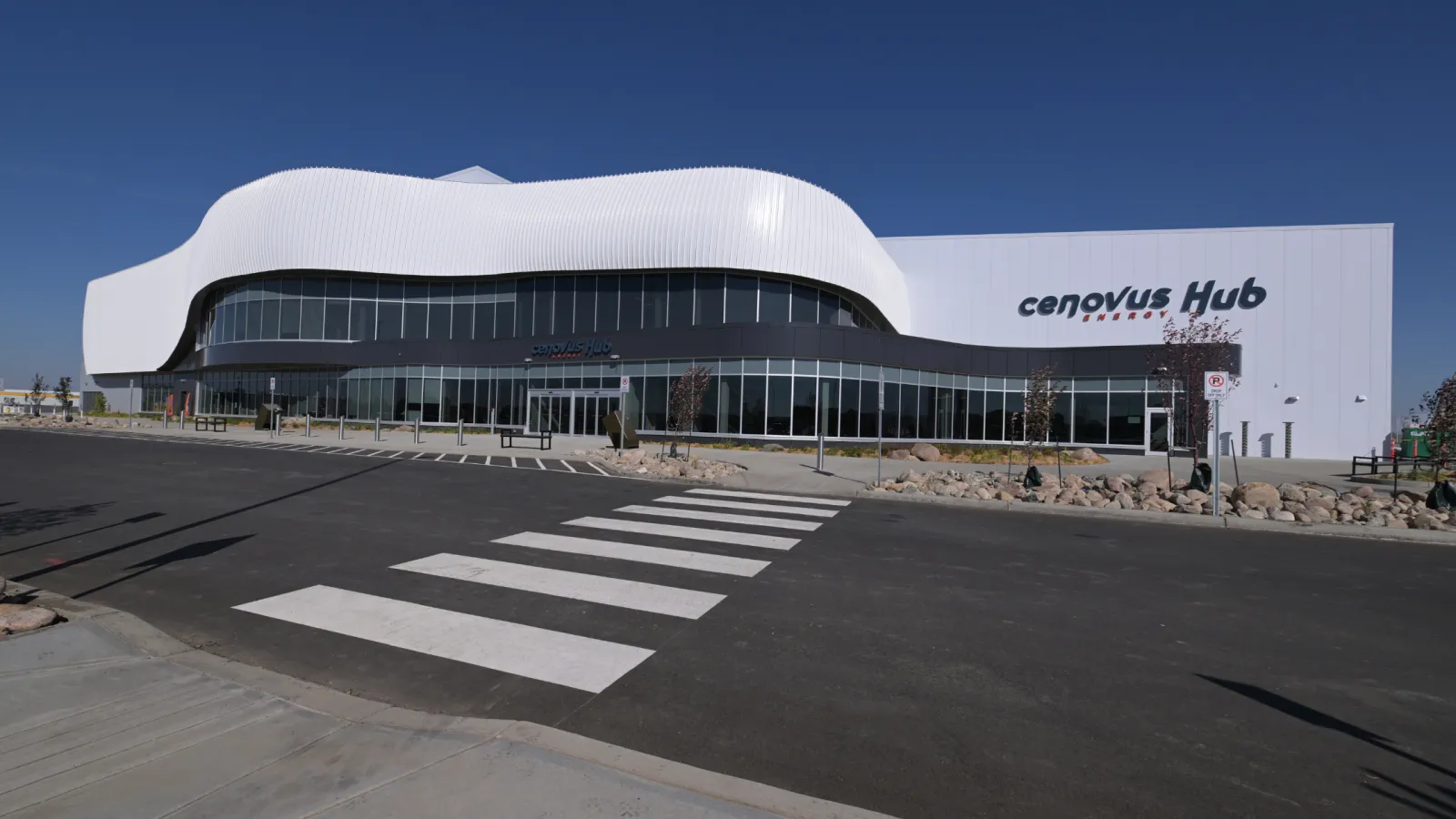 La entrada del Cenovus Energy Hub: una fachada superior blanca y curva sobre un vestíbulo con muros de vidrio, con el letrero cenovus Hub en el volumen de la arena y un paso peatonal en primer plano. — Cenovus Energy Hub