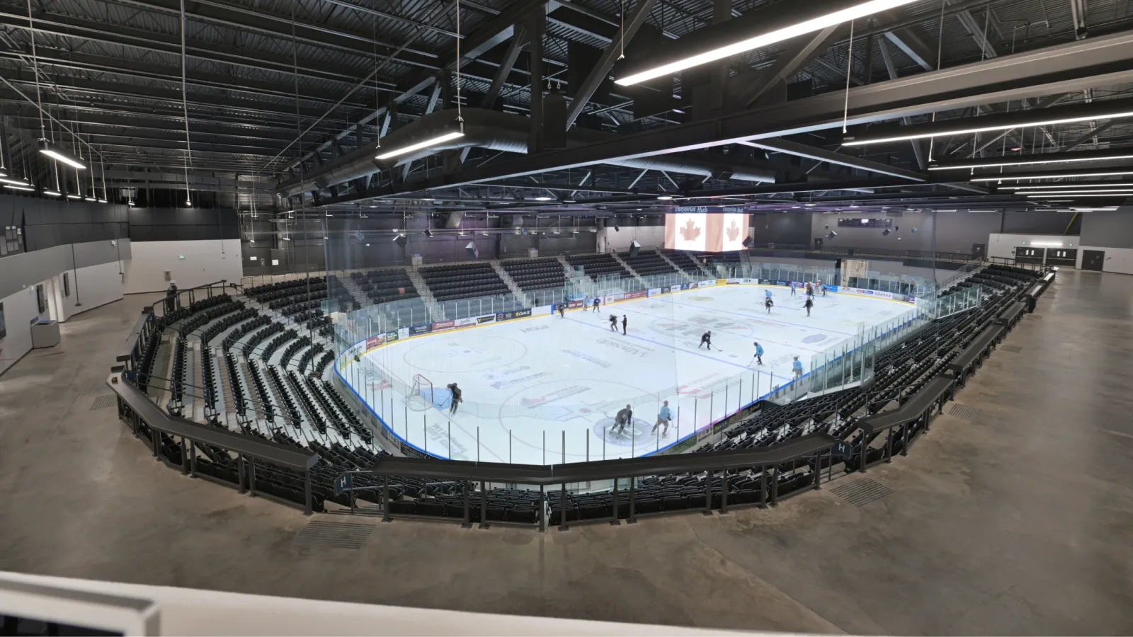 La arena de eventos desde el pasillo: toda la pista con bardas de vidrio y un marcador central colgante, rodeada de asientos oscuros, con algunos patinadores en el hielo. — Cenovus Energy Hub