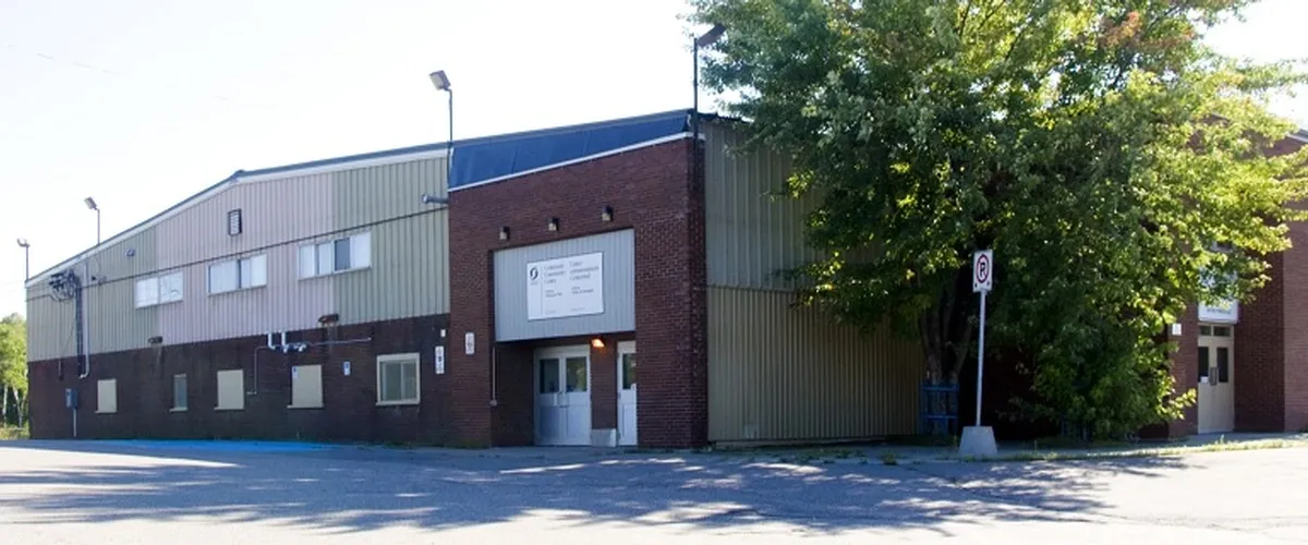 El edificio de la arena, de ladrillo y revestimiento metálico, con su entrada principal y el estacionamiento bajo los árboles. — Centennial Community Centre and Arena (Hanmer)