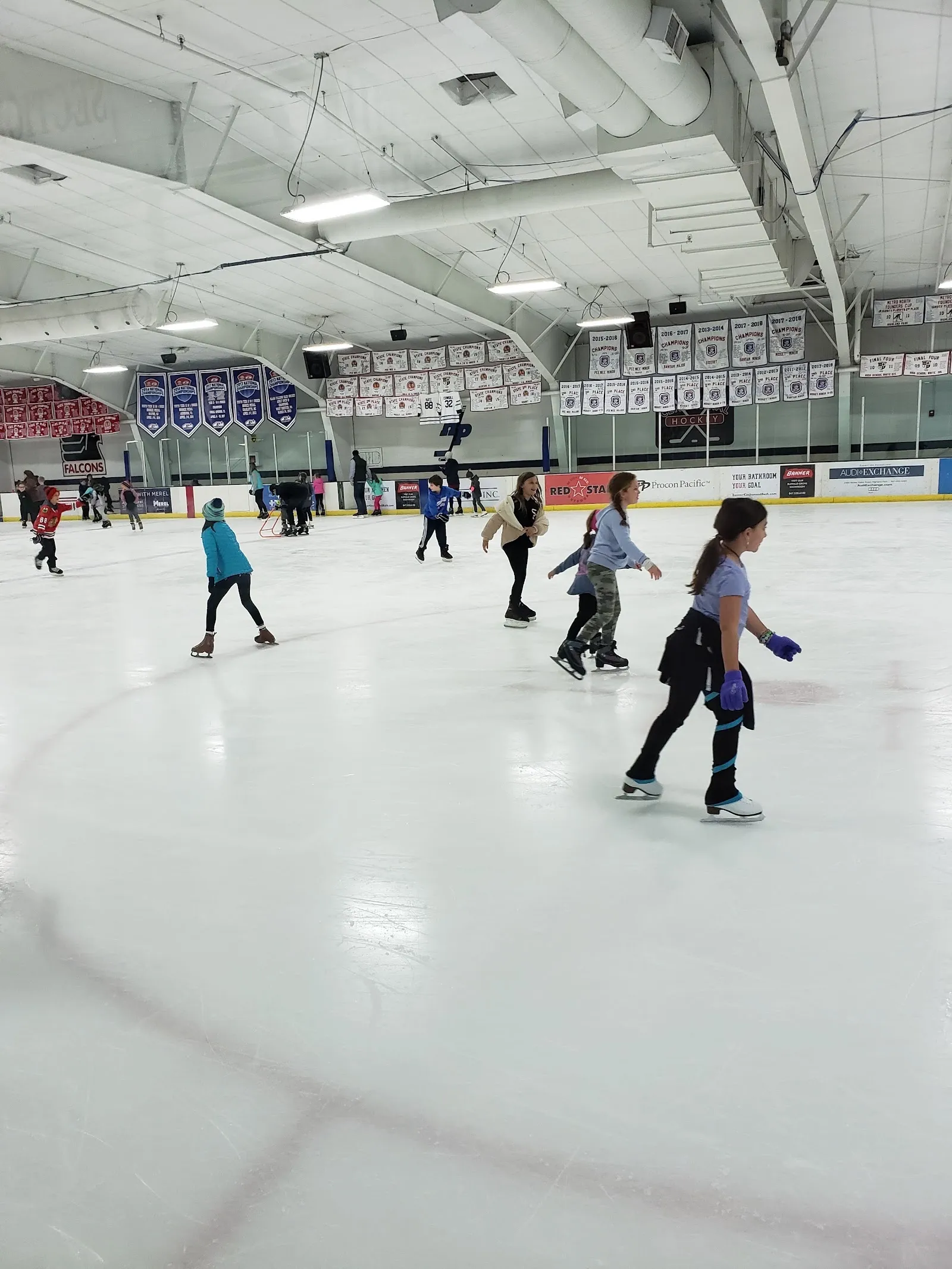 Patinadores repartidos por una pista blanca durante una sesión pública, vistos a través del vidrio, con un techo claro y banderines de la pista al fondo. — Centennial Ice Arena (Highland Park)