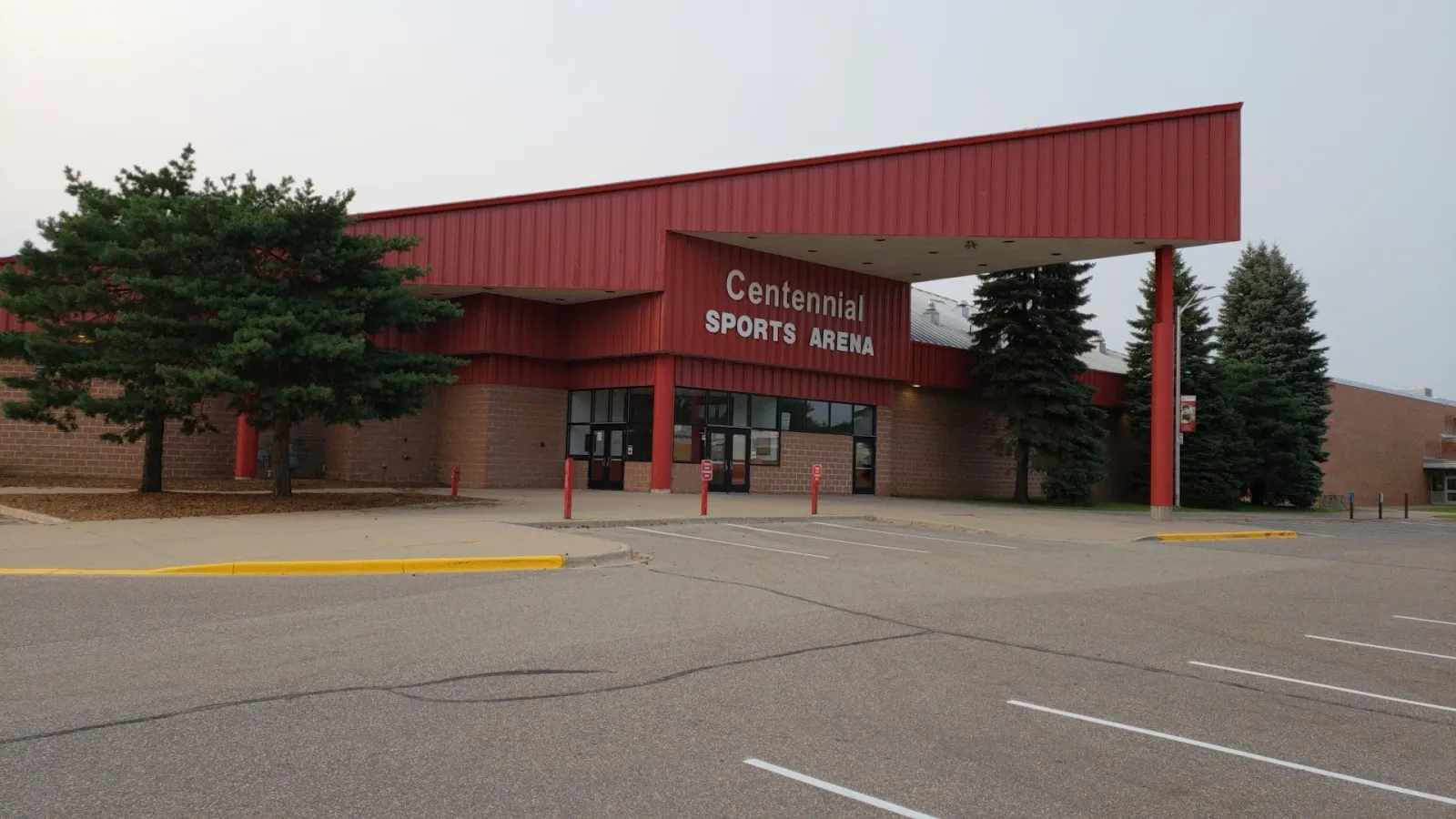 La entrada de ladrillo con la marquesina roja y el letrero 'Centennial Sports Arena', con abetos a ambos lados y el estacionamiento al frente. — Centennial Sports Arena