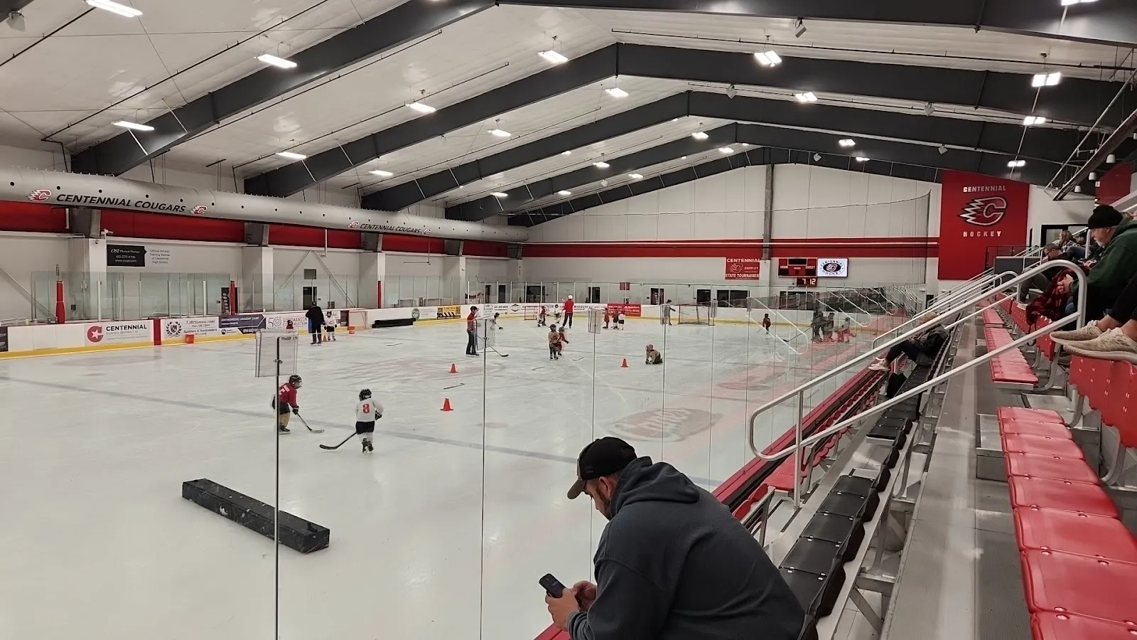 La pista vista desde los asientos rojos junto al vidrio durante un entrenamiento juvenil, con el logotipo de Centennial en la pared del fondo. — Centennial Sports Arena