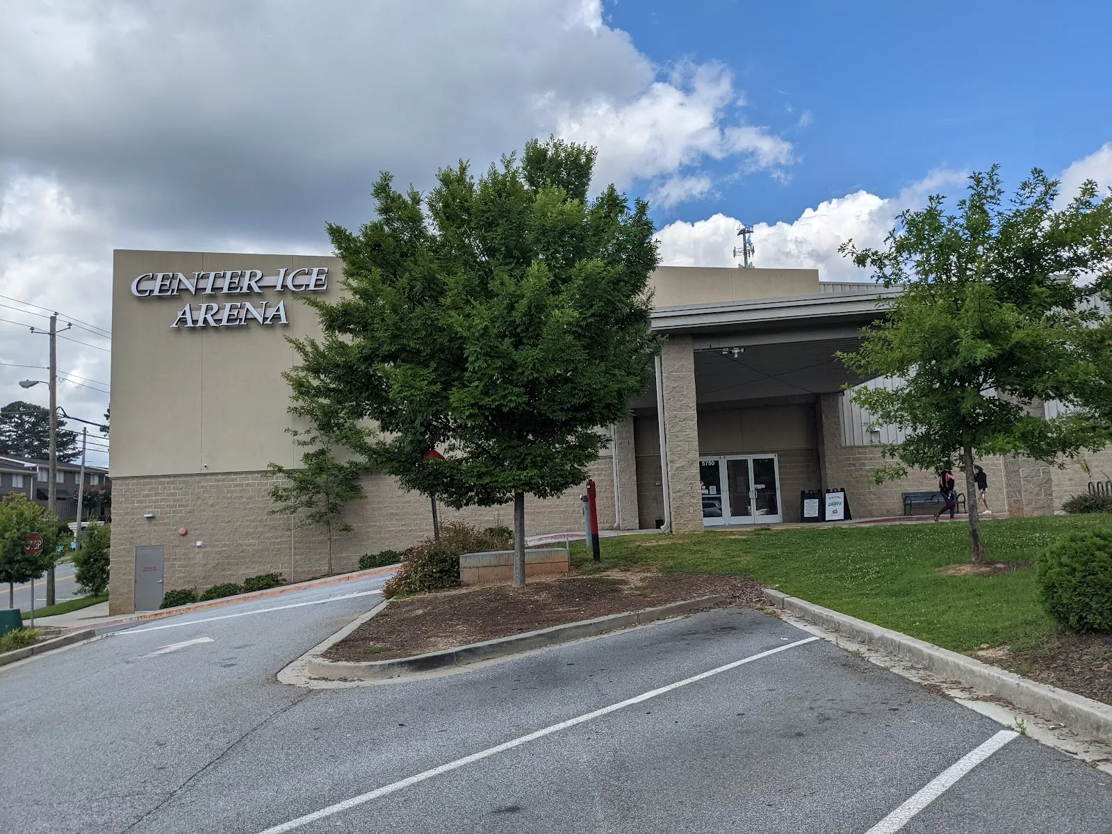 The tan block building with the Center Ice Arena sign and a covered entrance, seen across the parking lot past two young trees. — Center Ice Arena
