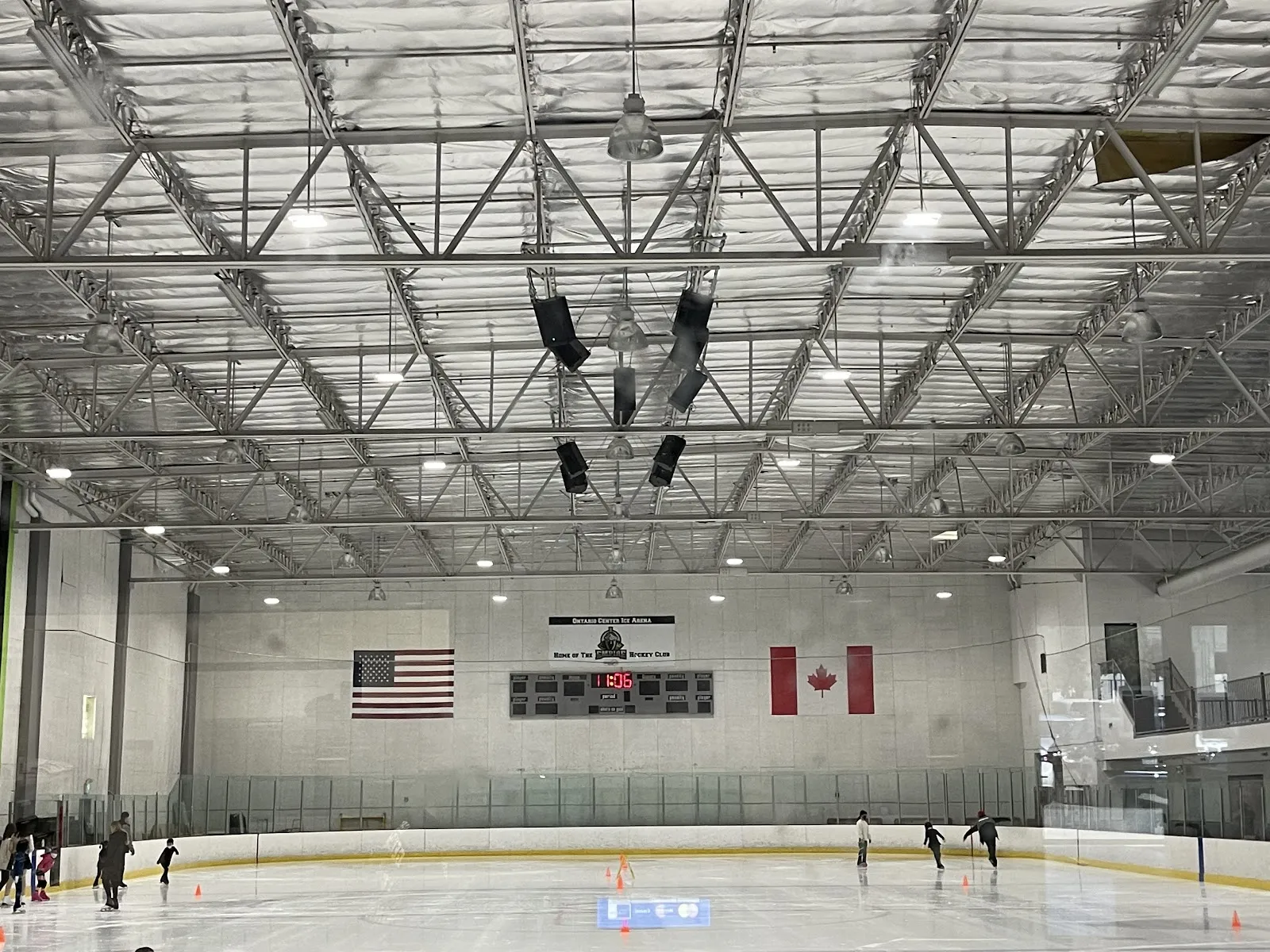 The sheet from one end under a steel-truss roof, with American and Canadian flags and a scoreboard on the far wall and a few skaters on the ice. — Ontario Center Ice Arena