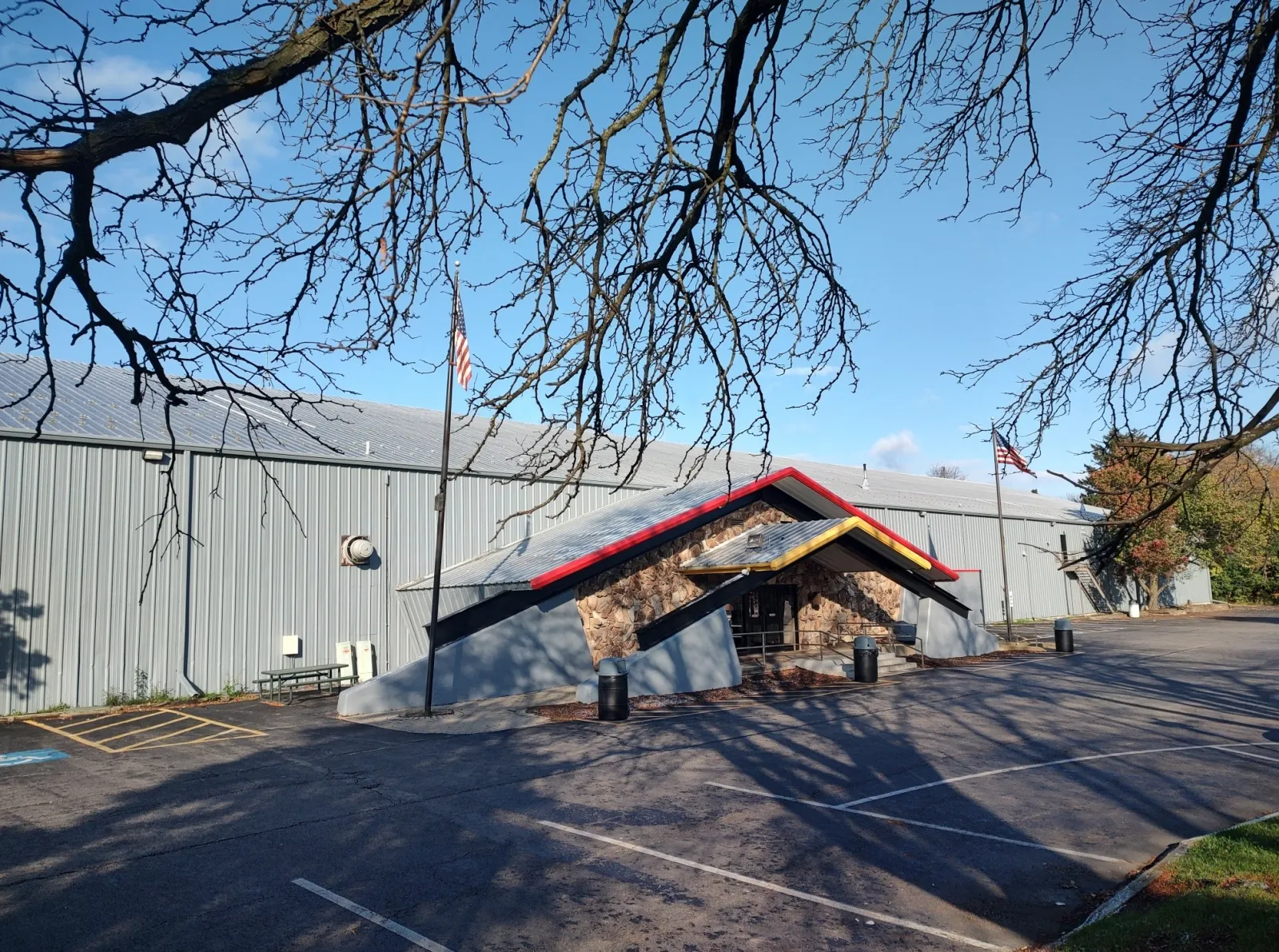 The rink's distinctive red-and-yellow A-frame stone entrance canopy in front of the grey metal-sided arena building, parking lot in foreground. — Center Ice of DuPage