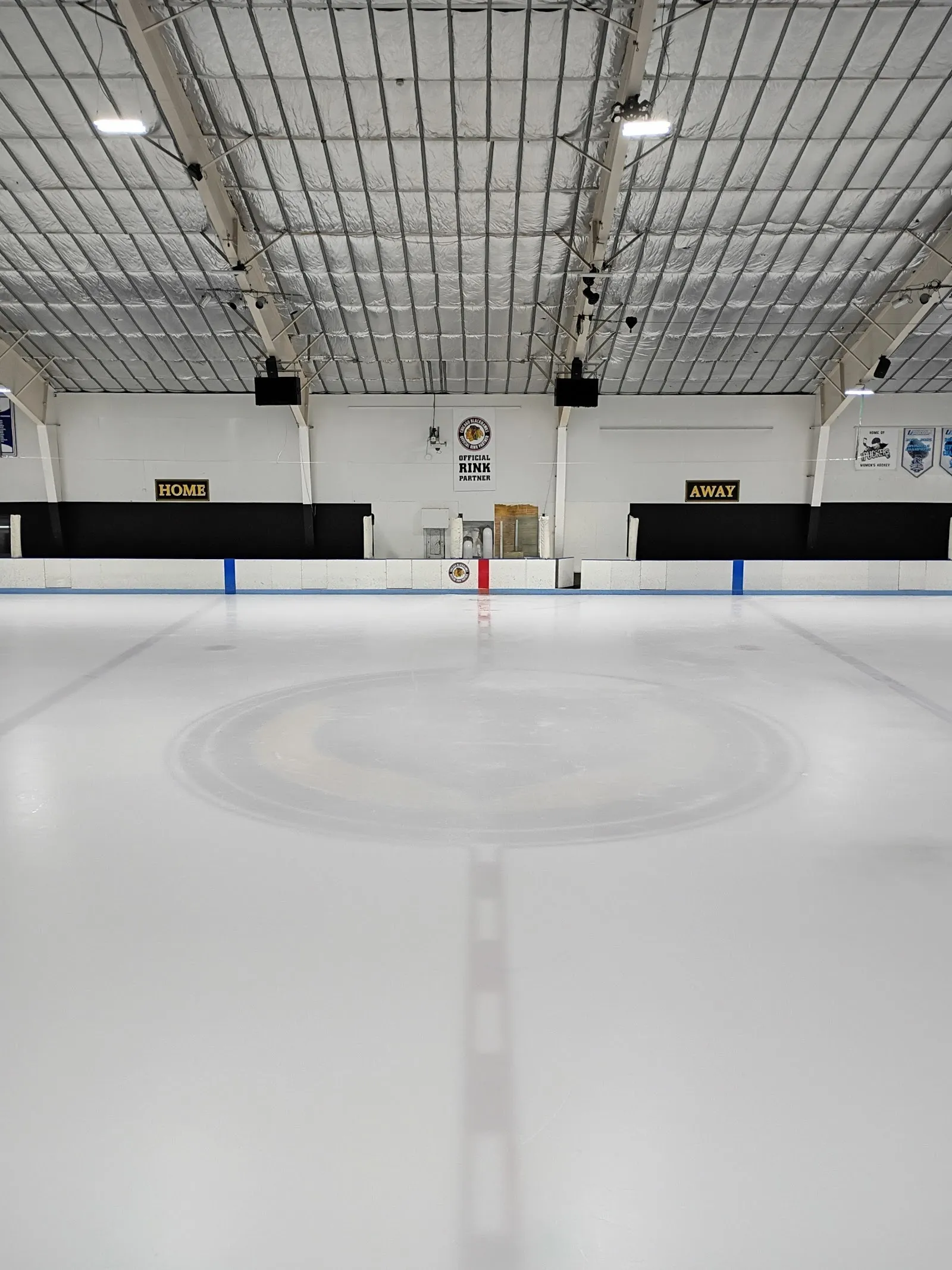 An empty sheet with a faint centre circle seen from the end boards, a ceiling of steel trusses and black boards along the far wall. — Center Ice of DuPage