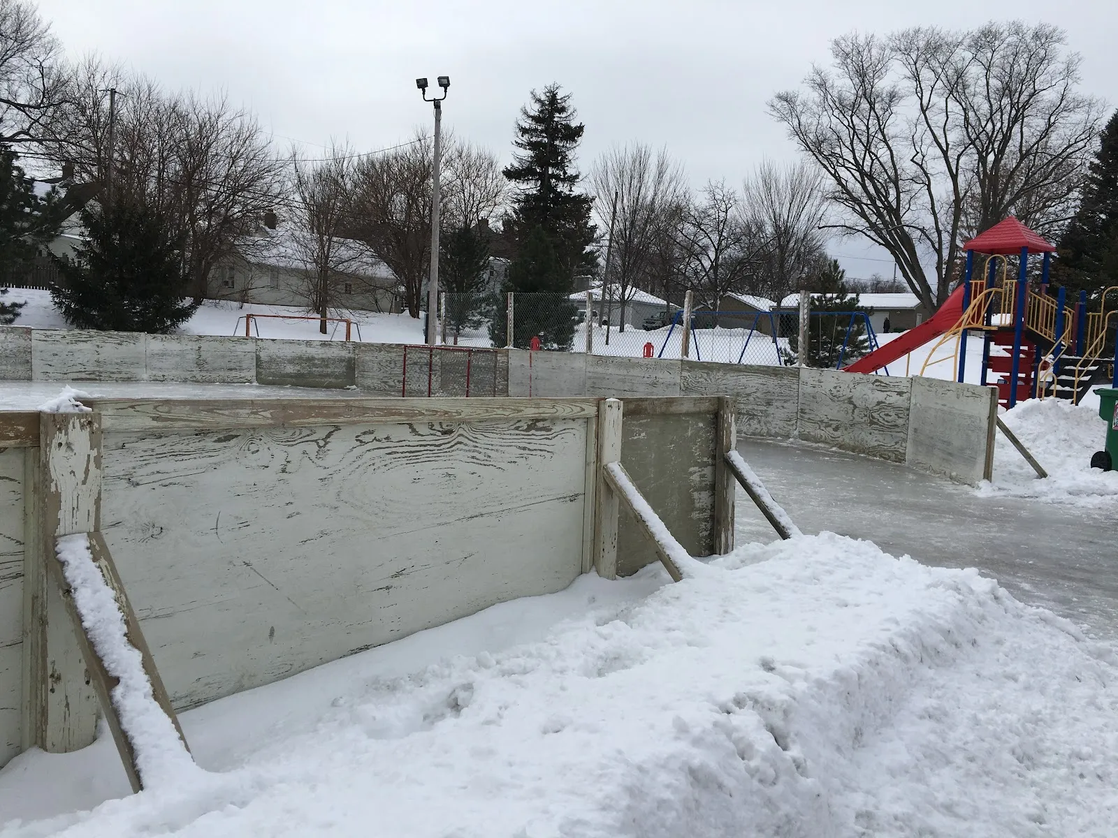 The snow-banked board wall of the outdoor rink at Center Park, with the playground beyond — Center Park Outdoor Rink