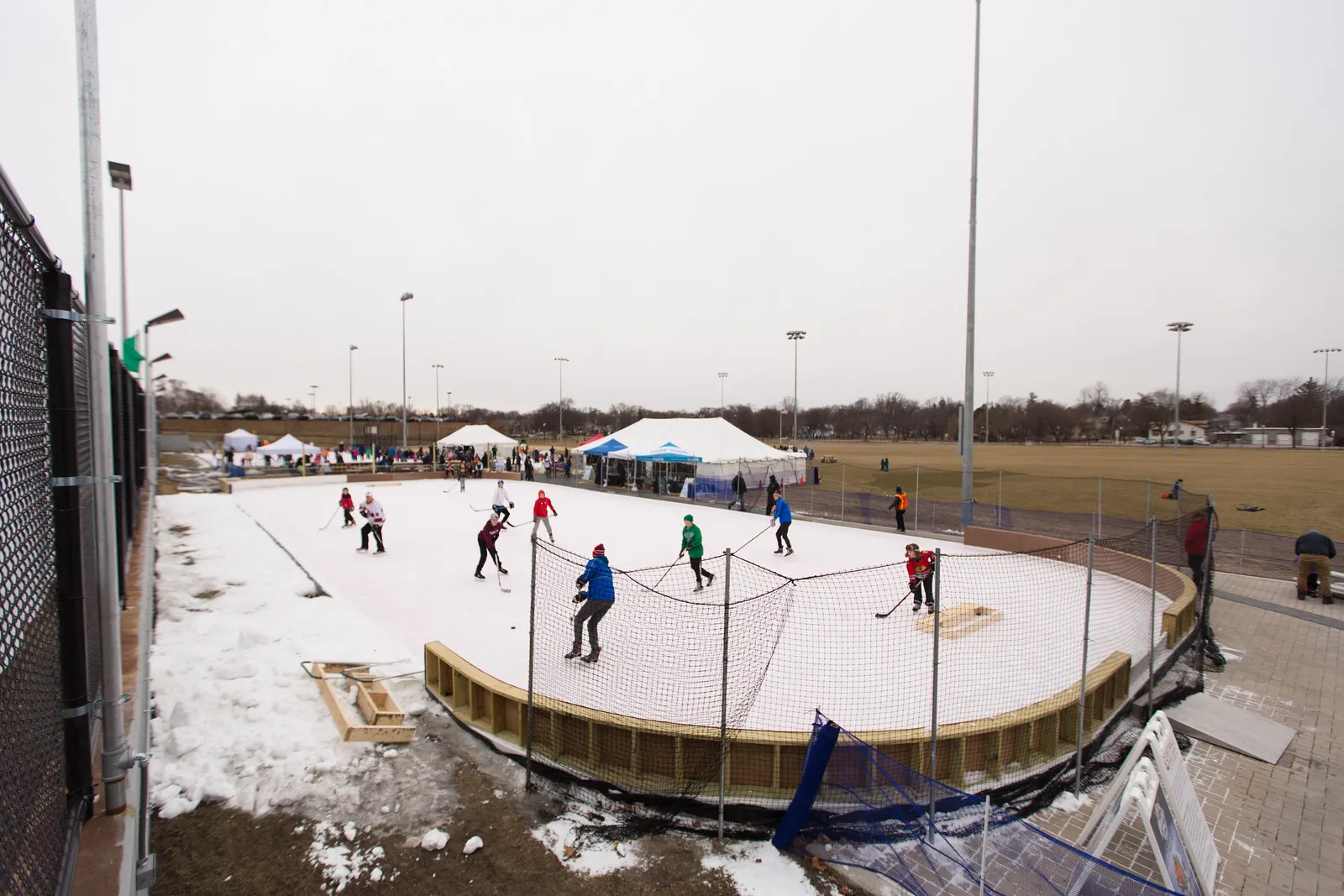 A boarded outdoor rink with netting and floodlights at the Central Athletic Complex during a hockey event, snow still on the ground beside it. — Central Athletic Complex Ice Rinks (Wheaton)