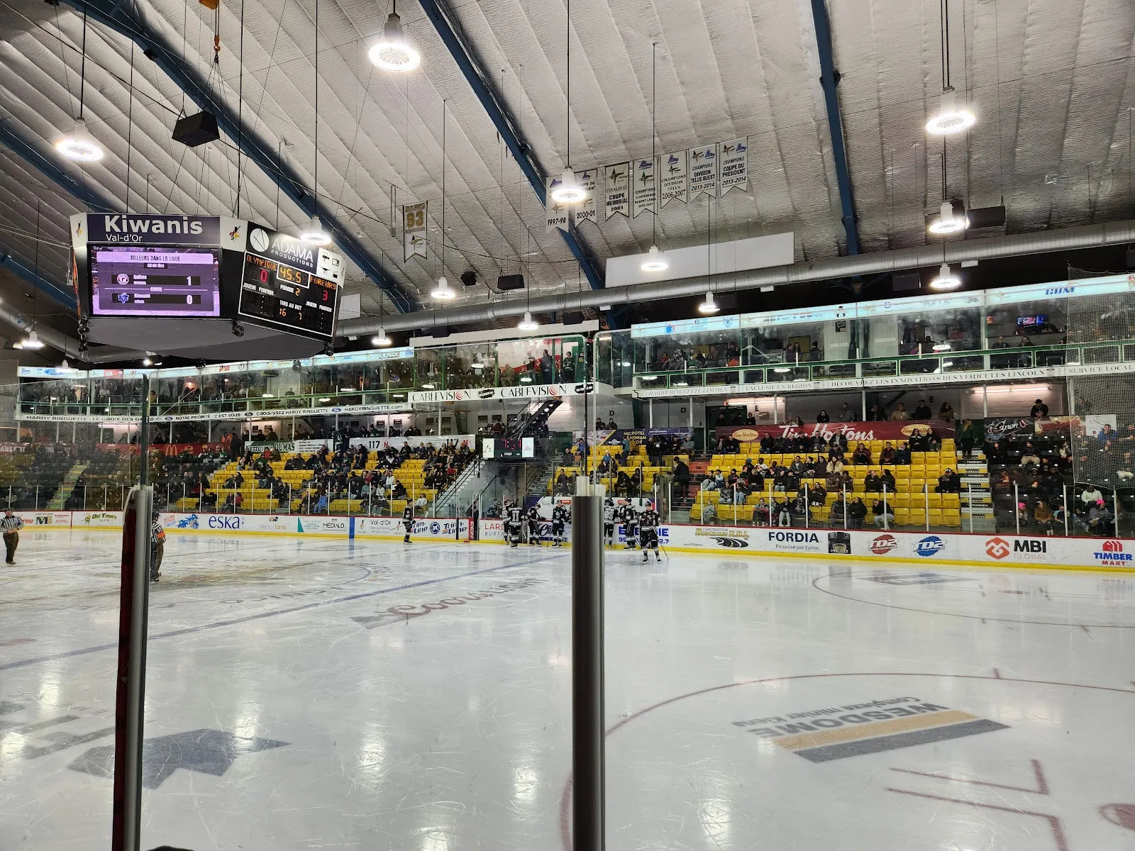 Une partie en cours sur la glace de l’Aréna Kiwanis de Val-d’Or, avec le nom Kiwanis sur le tableau indicateur et des spectateurs dans les gradins derrière la baie vitrée. — Centre Agnico Eagle et Aréna Kiwanis