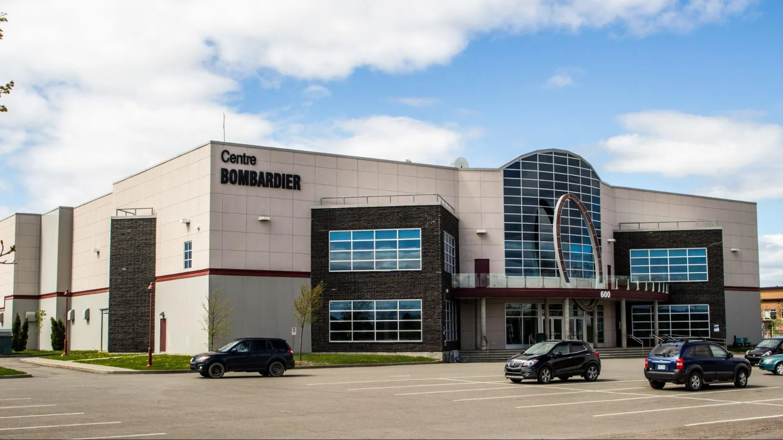 Le Centre Bombardier à La Pocatière : un bâtiment moderne en verre et panneaux gris, son nom au-dessus de l'entrée et un stationnement devant. — Centre Bombardier