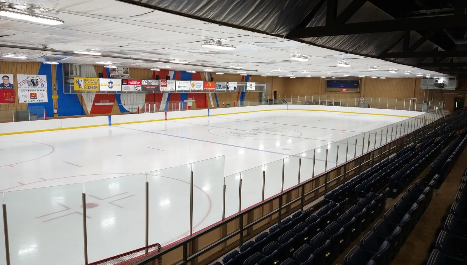 La patinoire vue du haut des sièges bleus, avec des bannières de commanditaires et des panneaux muraux rouges et bleus au fond, sous un toit blanc. — Centre communautaire Michel-Labadie