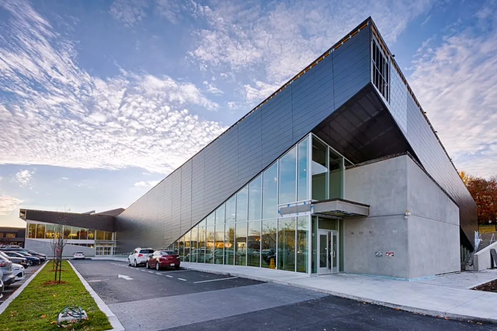 Le bâtiment anguleux revêtu de gris, avec son angle vitré et son entrée, vu depuis le stationnement dans la lumière du soir. — Centre de la culture et du sport de Sainte-Julie