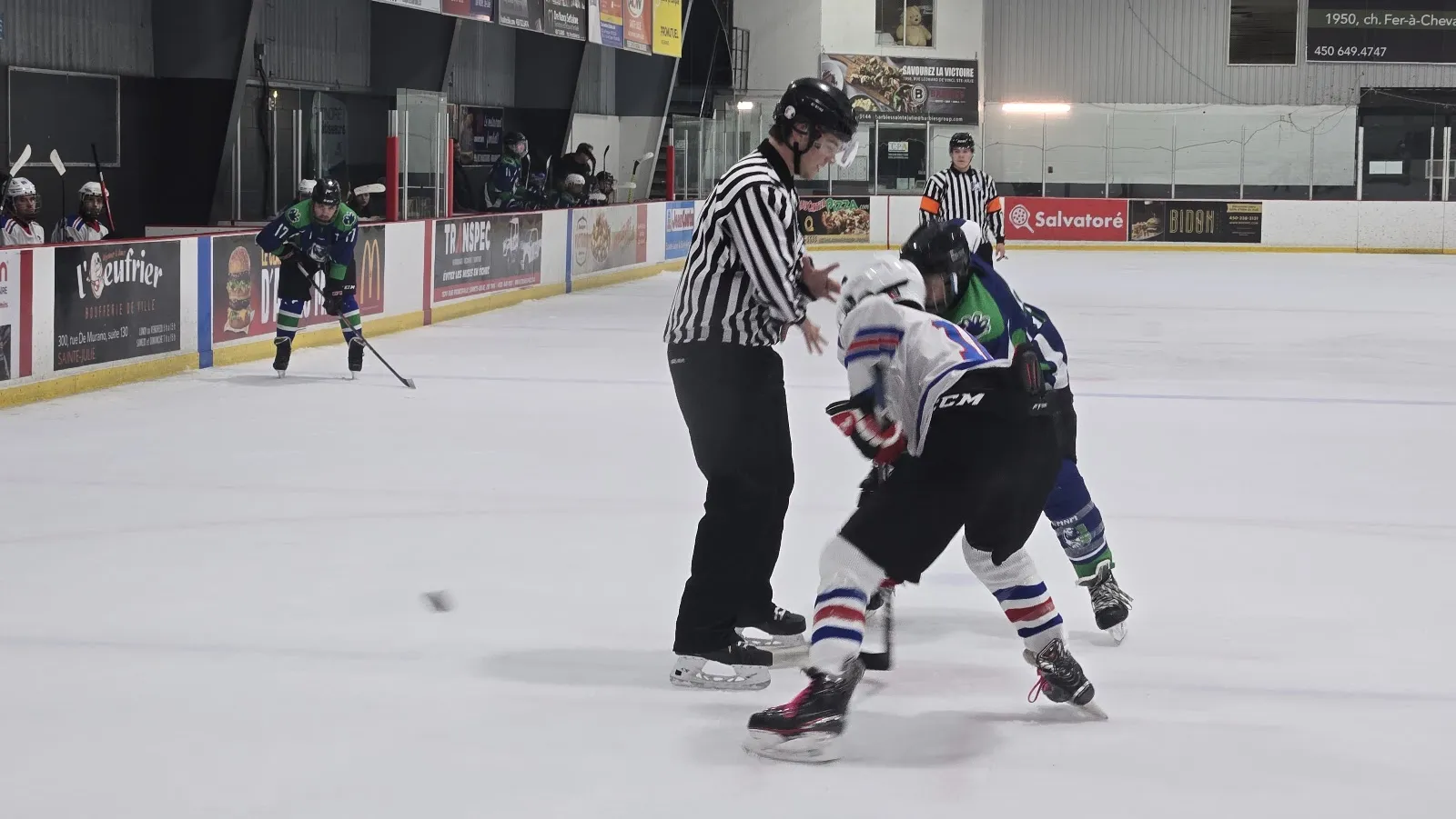 Une mise au jeu pendant un match de hockey sur la glace de l'aréna du Centre de la culture et du sport de Sainte-Julie : un arbitre laisse tomber la rondelle entre deux joueurs, avec des panneaux de commanditaires le long de la bande et les bancs derrière. — Centre de la culture et du sport de Sainte-Julie