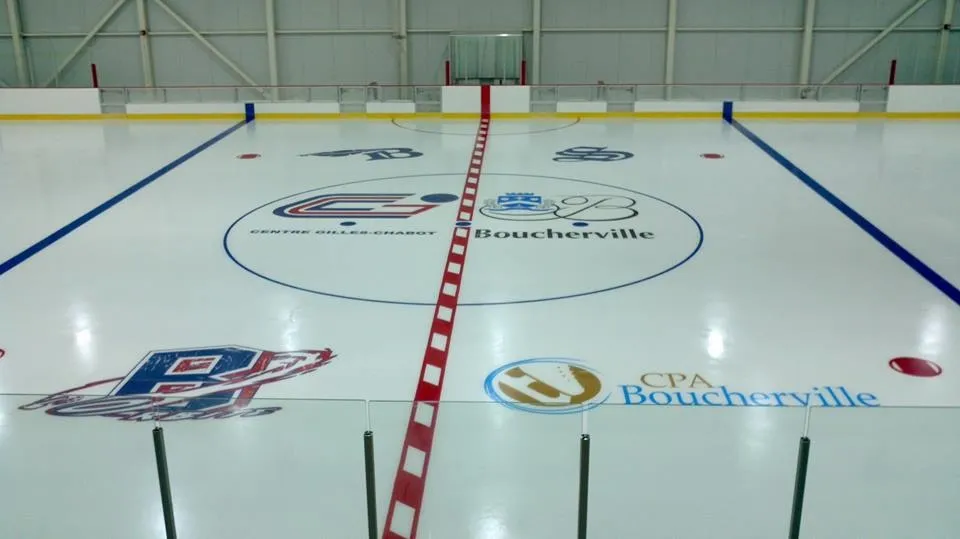 A wide empty sheet seen from the end, the blue lines and a red centre line across white ice with a Boucherville logo at centre circle. — Centre des glaces Gilles-Chabot