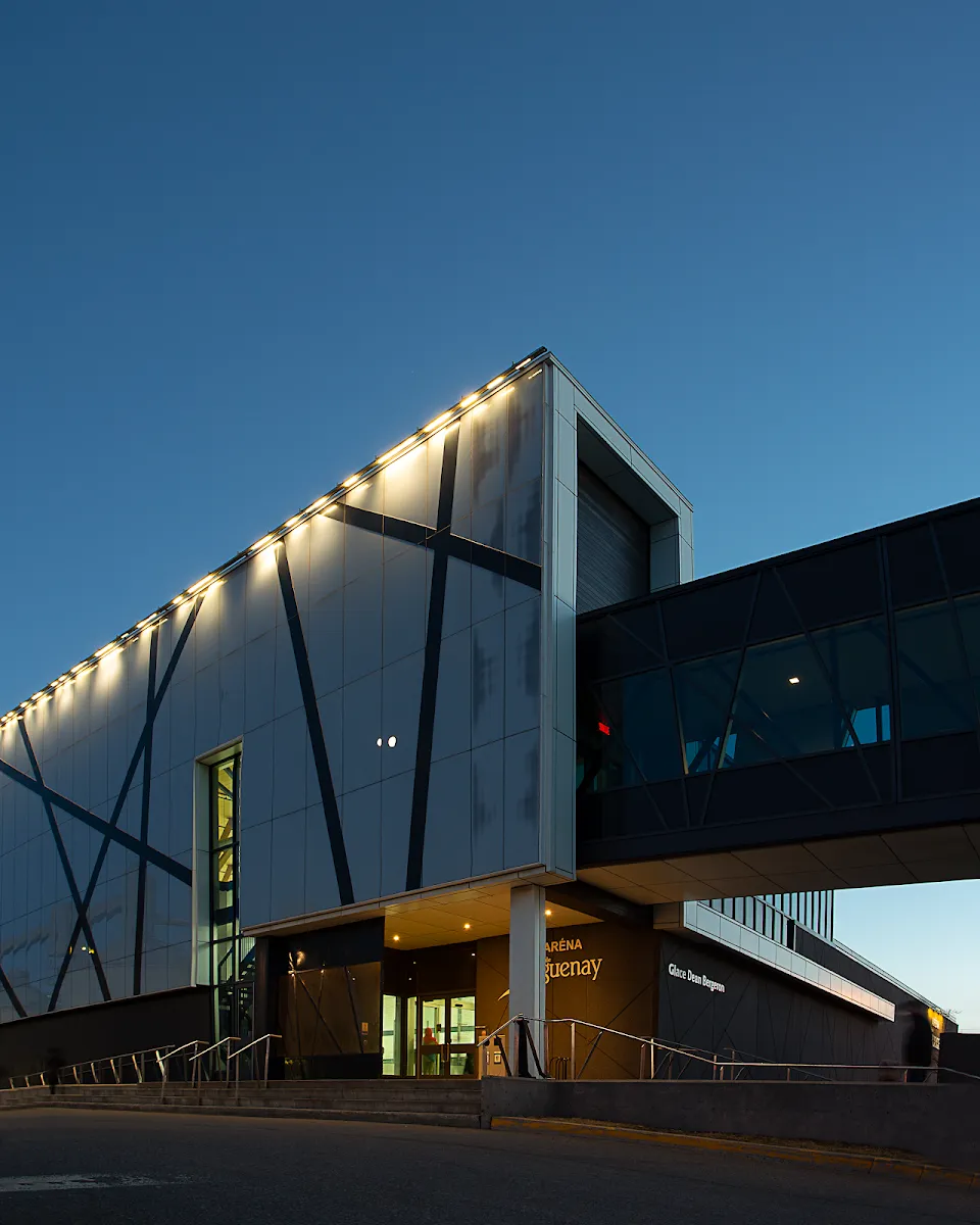 The lit glass-and-metal facade and entrance of the arena building at the Centre des sports Jean-Claude-Tremblay in La Baie at dusk — Centre des sports Jean-Claude-Tremblay