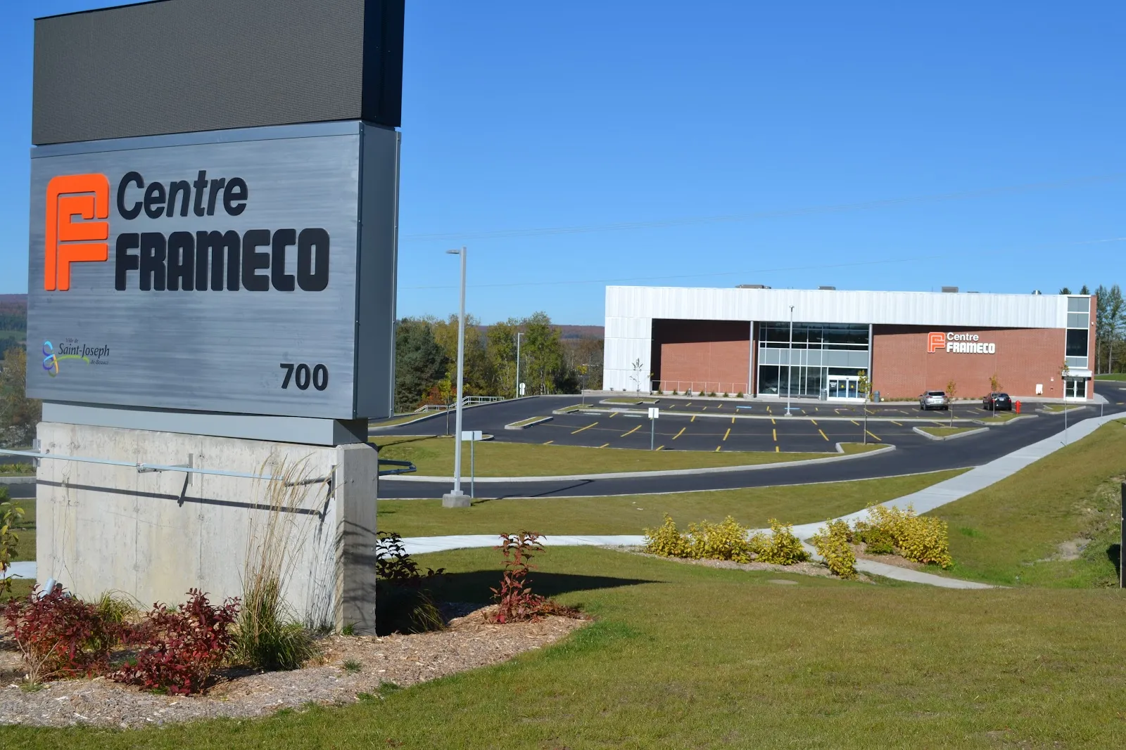 The Centre Frameco entrance sign in the foreground and, past a sloping lawn and the parking lot, the long building with its white metal upper wall, red brick base and glass entrance, under a clear sky. — Centre Frameco