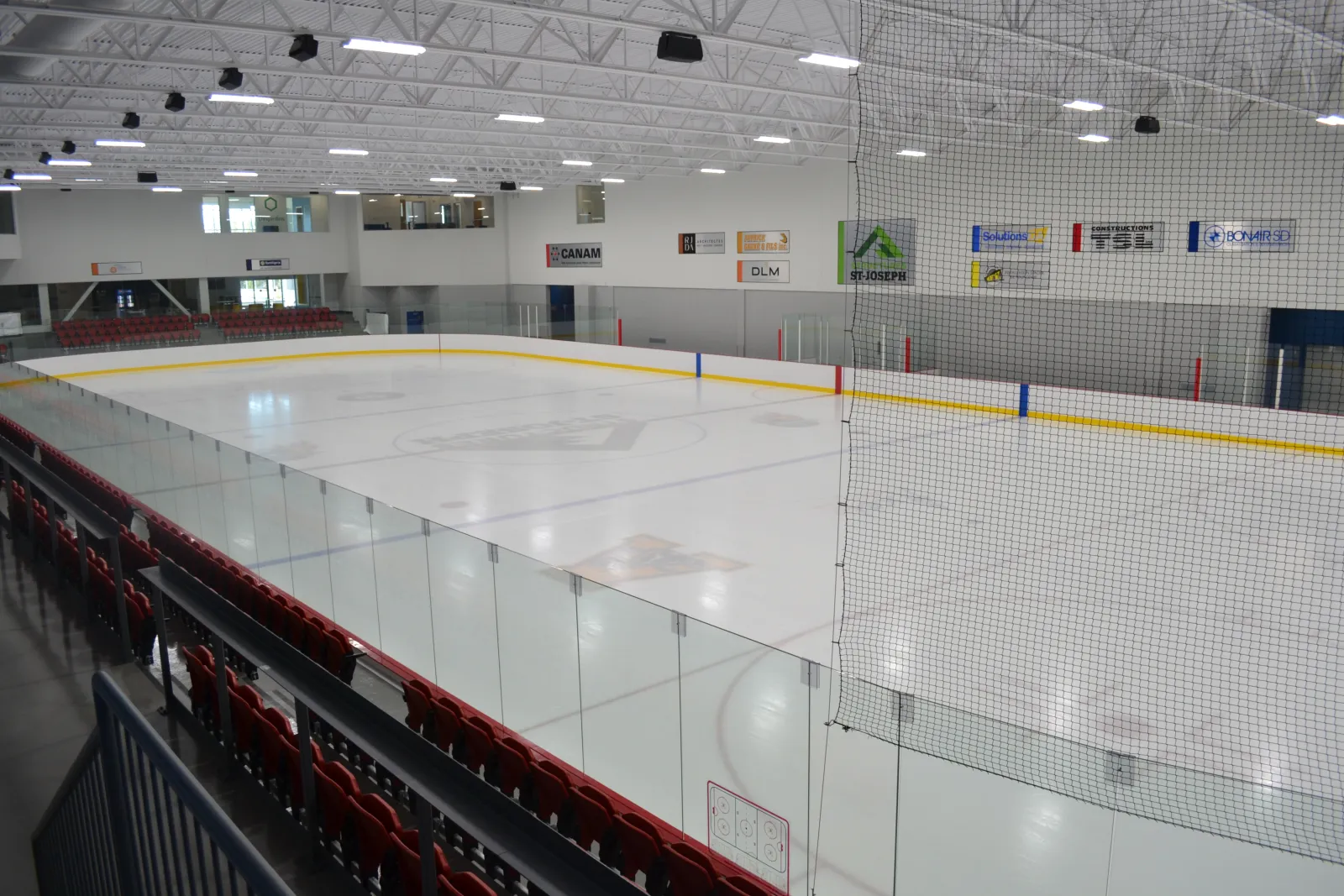 The empty ice sheet seen from a corner of the stands, with red seats along the glass, a safety net over the end boards and sponsor signs on the far wall below the upper windows. — Centre Frameco