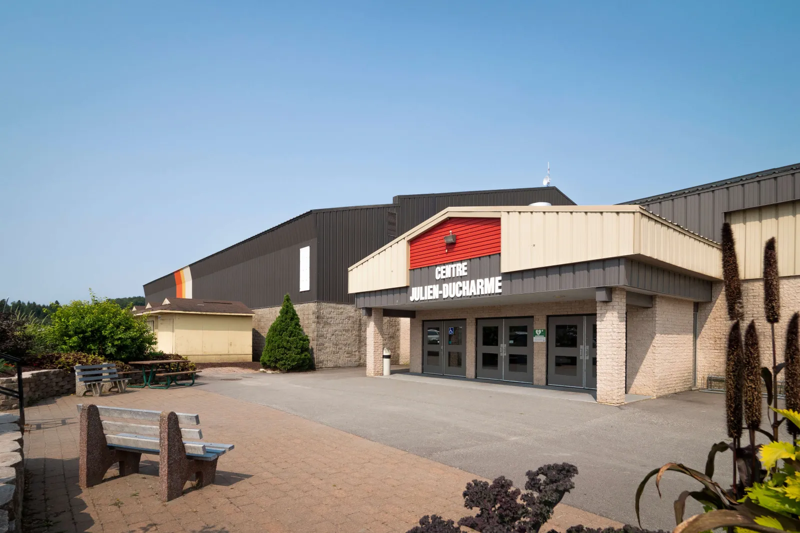 Un volume d’entrée beige et gris portant l’enseigne « Centre Julien-Ducharme » sous une bande rouge, avec la salle sombre de l’aréna derrière et des bancs sur une place pavée. — Centre Julien-Ducharme