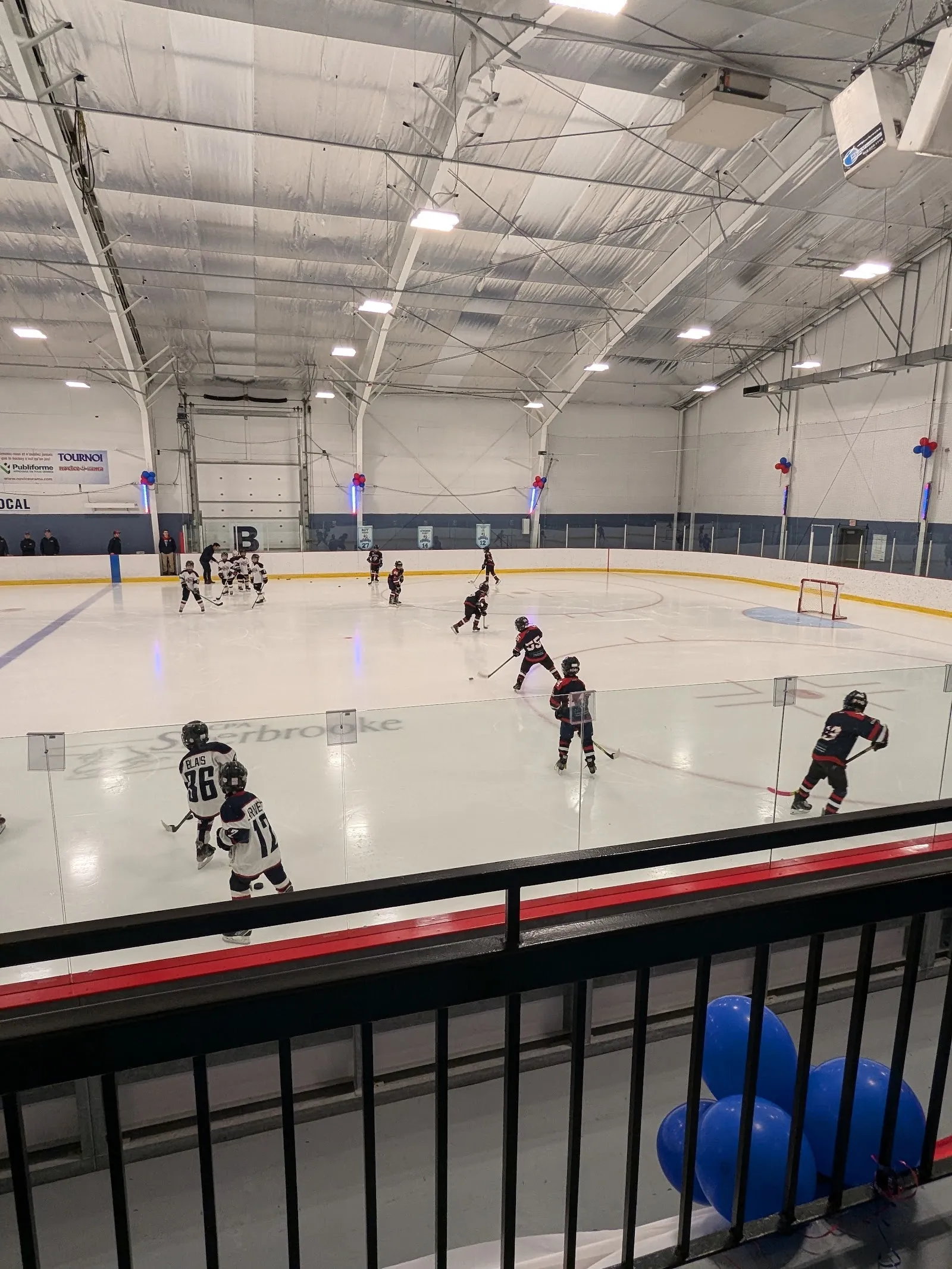 Un match sur une grande surface de glace vu de la rampe, des joueurs répartis sur la glace sous un haut plafond à charpente d’acier avec une bande de fenêtres. — Centre Julien-Ducharme