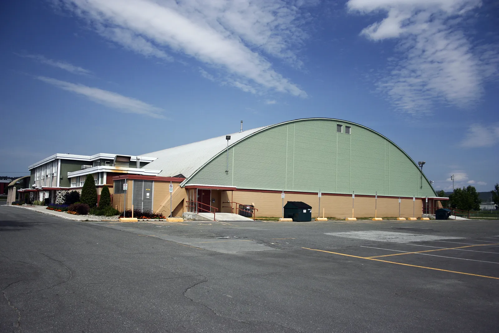 Le long aréna vert à toit en arche, à côté de son bloc d’entrée de deux étages et d’un grand stationnement, sous un ciel bleu. — Centre Mario-Gosselin
