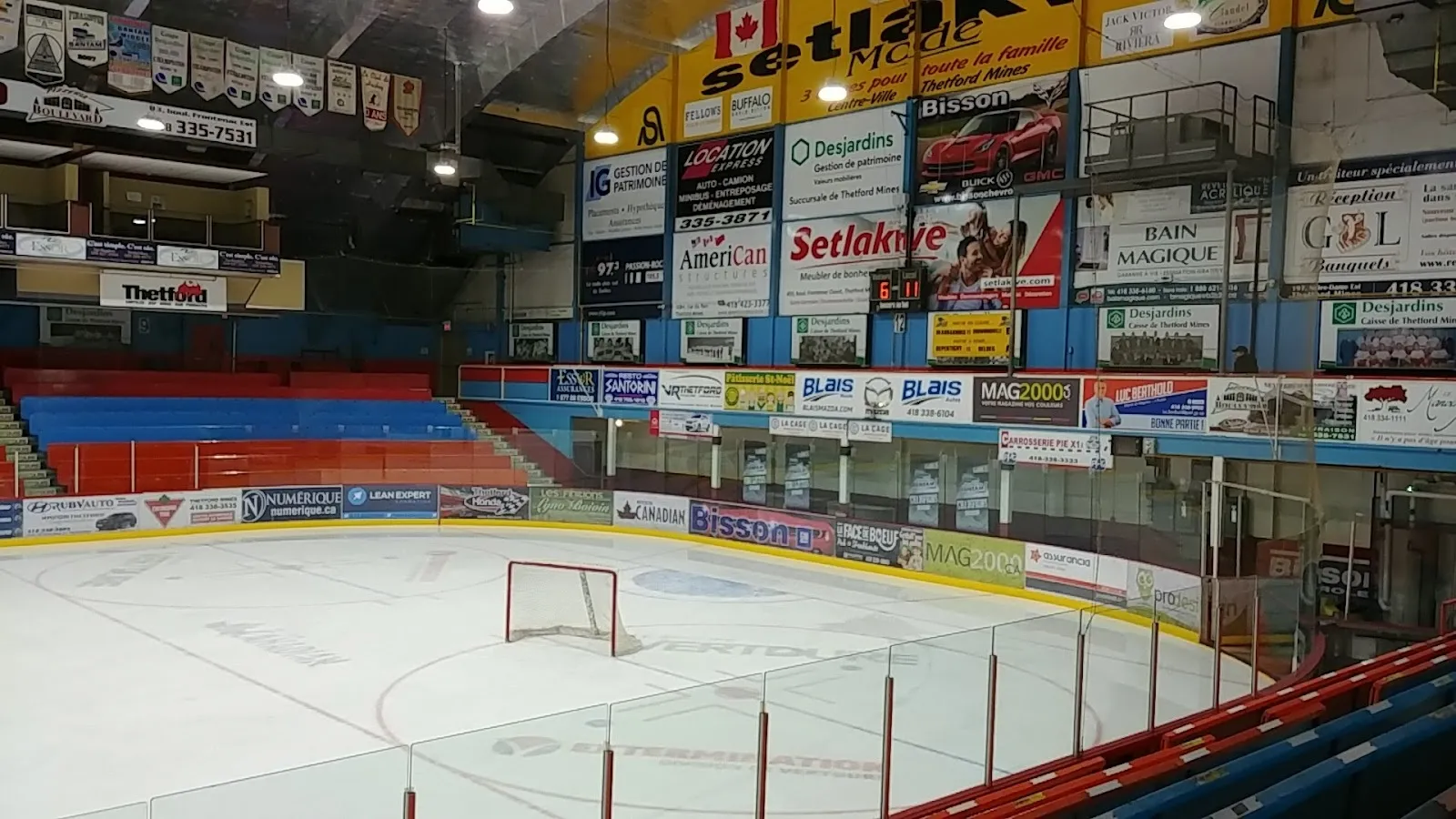 Une vue large de l’aréna depuis les gradins, une glace vide et un but, avec des panneaux de commanditaires et des bannières au-dessus des sièges qui l’entourent. — Centre Mario-Gosselin