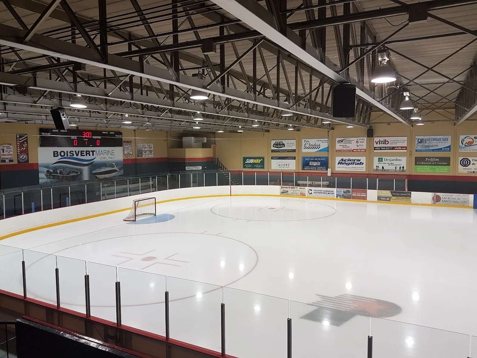 The ice surface seen from the stands of the Aussant arena, with the boards and sponsor signs and the steel roof trusses — Centre récréatif Aussant (Aréna)
