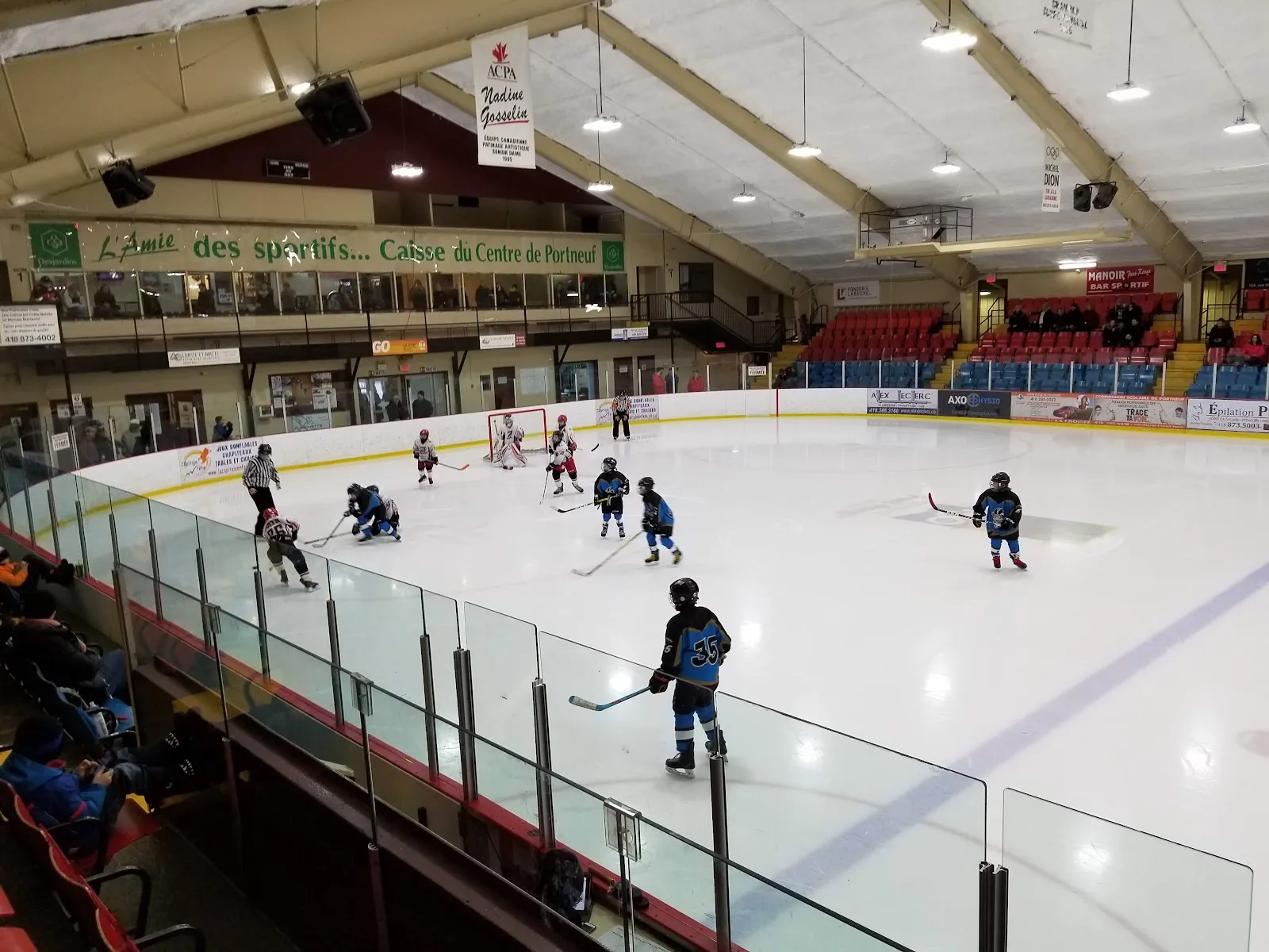A children's hockey practice on the ice of the Centre récréatif Joé-Juneau, seen from the glass with the stands and banners behind — Centre récréatif Joé-Juneau