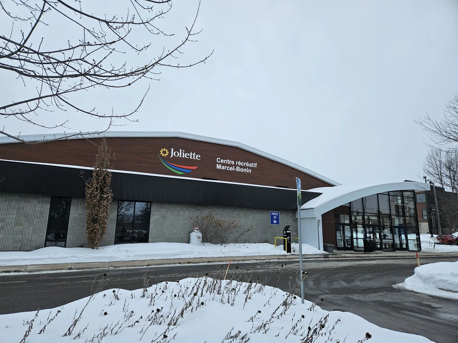 The low brick and dark-panel front of the arena in winter, signed Joliette Centre récréatif Marcel-Bonin, with a curved glass entrance at right. — Centre récréatif Marcel-Bonin