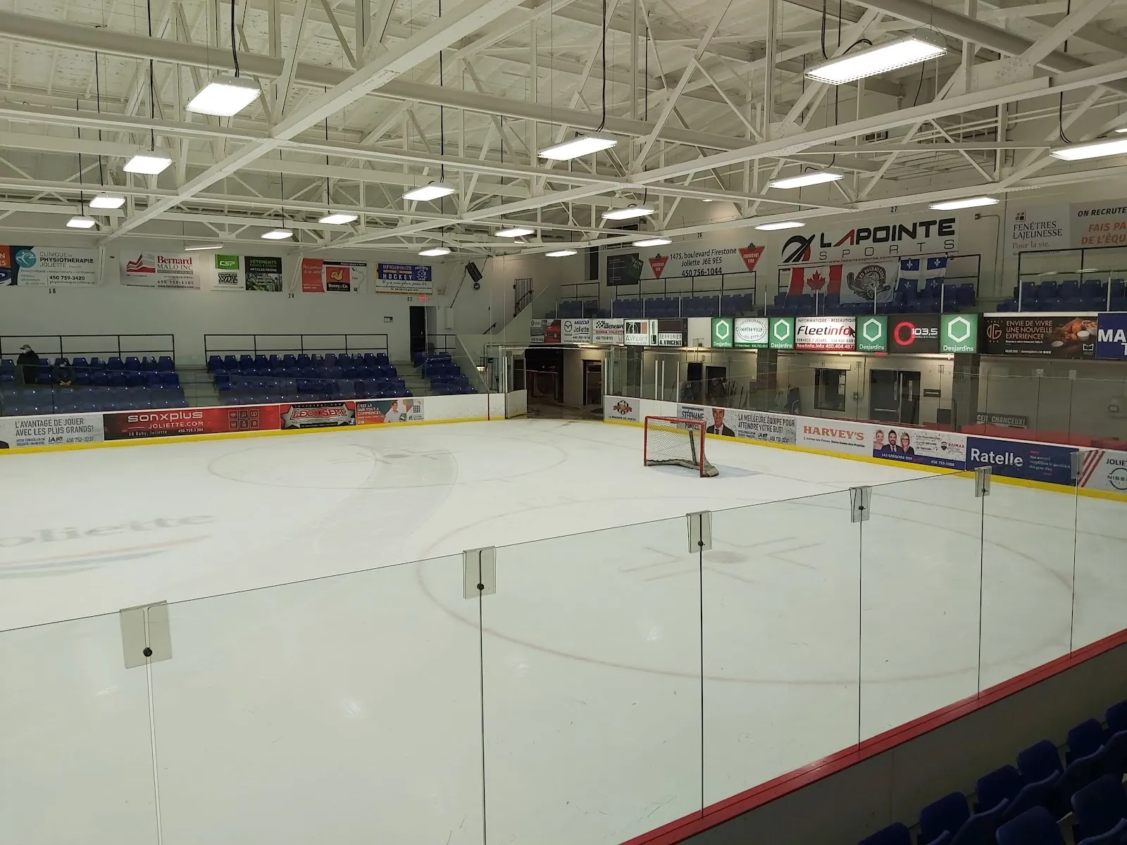 The empty rink from the glass at one end, with blue seats along the far side and sponsor boards under a white steel roof. — Centre récréatif Marcel-Bonin