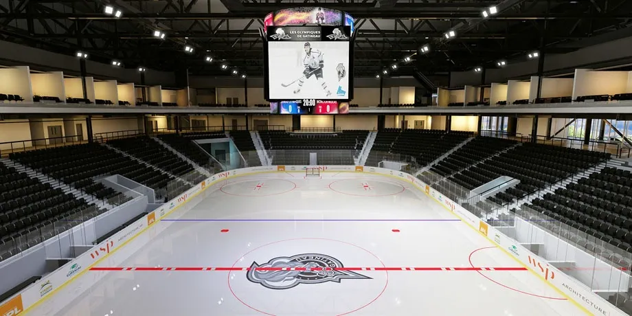 The empty arena bowl seen from the end, a long sheet with a red centre line and a Slush Puppie logo at centre under a steel roof and dark seating. — Centre Slush Puppie