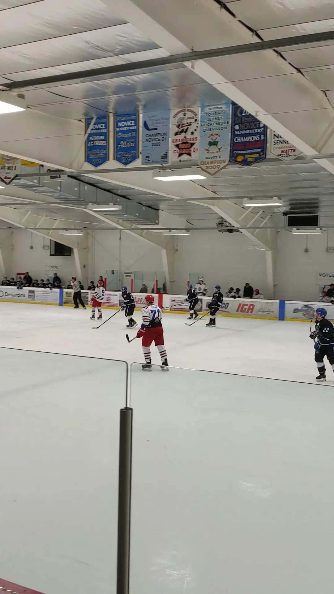 A hockey game on the ice at the Centre sportif André-Dubé in Senneterre, with rows of championship banners hanging from the white roof trusses. — Centre sportif André-Dubé