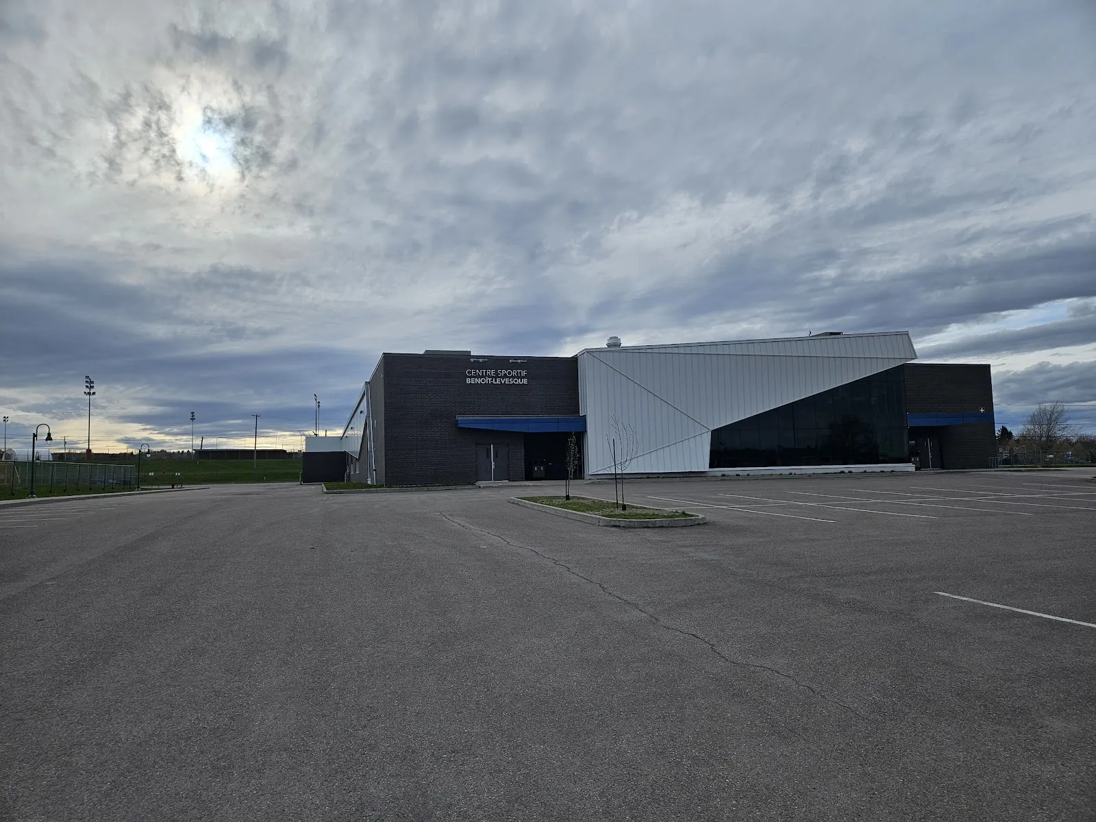 El Centre sportif Benoît-Levesque visto desde el otro lado de su estacionamiento: un bloque oscuro con un ala blanca en ángulo y marquesinas azules. — Centre sportif Benoît-Levesque