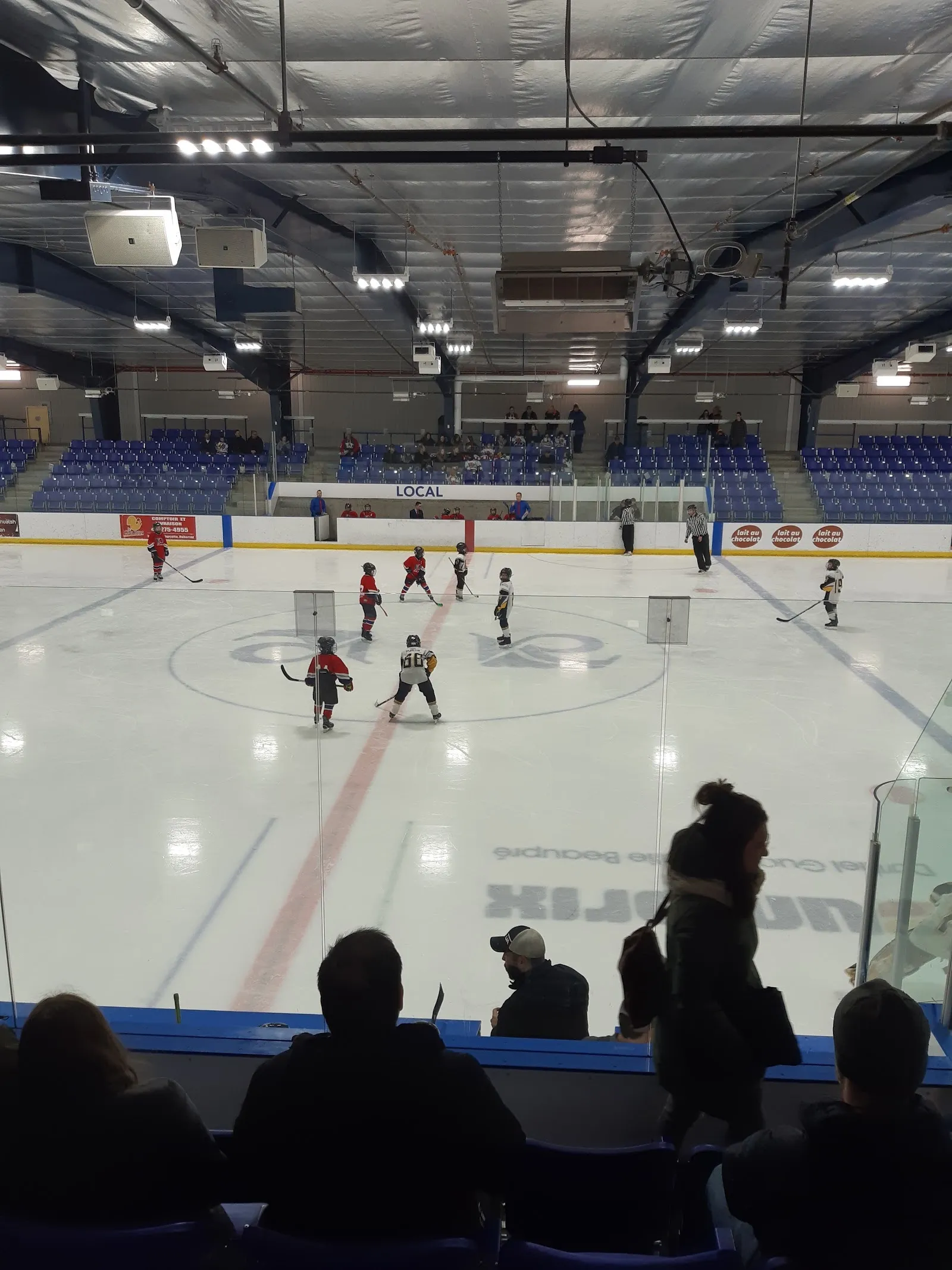 Un partido visto desde las gradas, con jugadores repartidos por una pista luminosa bajo un alto techo con una pared de marcadores en el extremo opuesto. — Centre sportif Benoît-Levesque