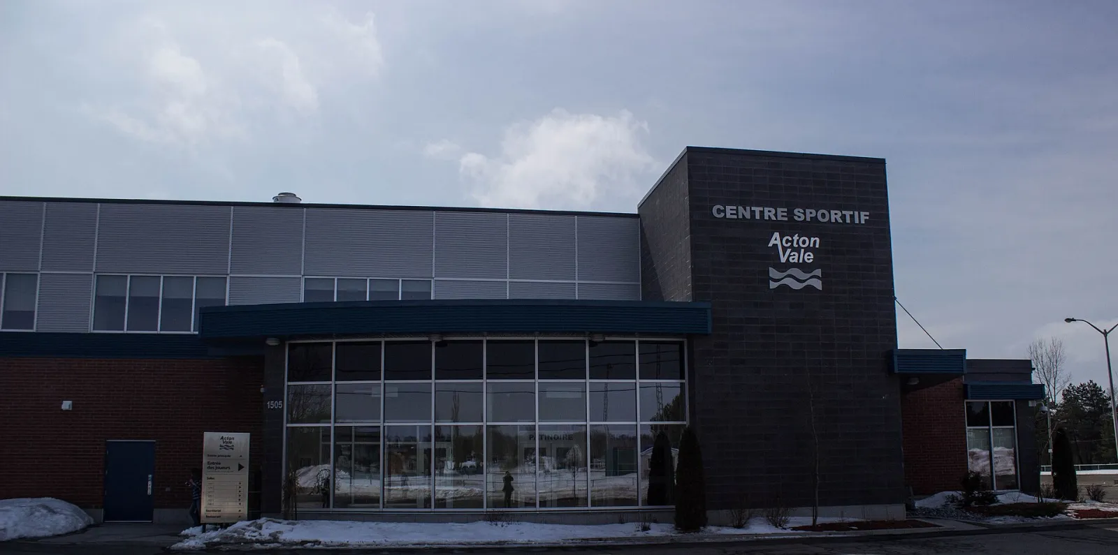 El Centre sportif visto desde el lado del estacionamiento: un frente de ladrillo y vidrio de dos pisos con una torre oscura que lleva el letrero del Centre sportif Acton Vale. — Centre sportif d'Acton Vale