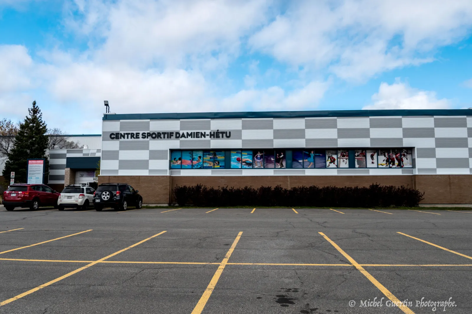 The Centre sportif Damien-Hétu seen across its car park, with the name in black letters above a window band of swimming, skating and hockey images. — Centre sportif Damien-Hétu