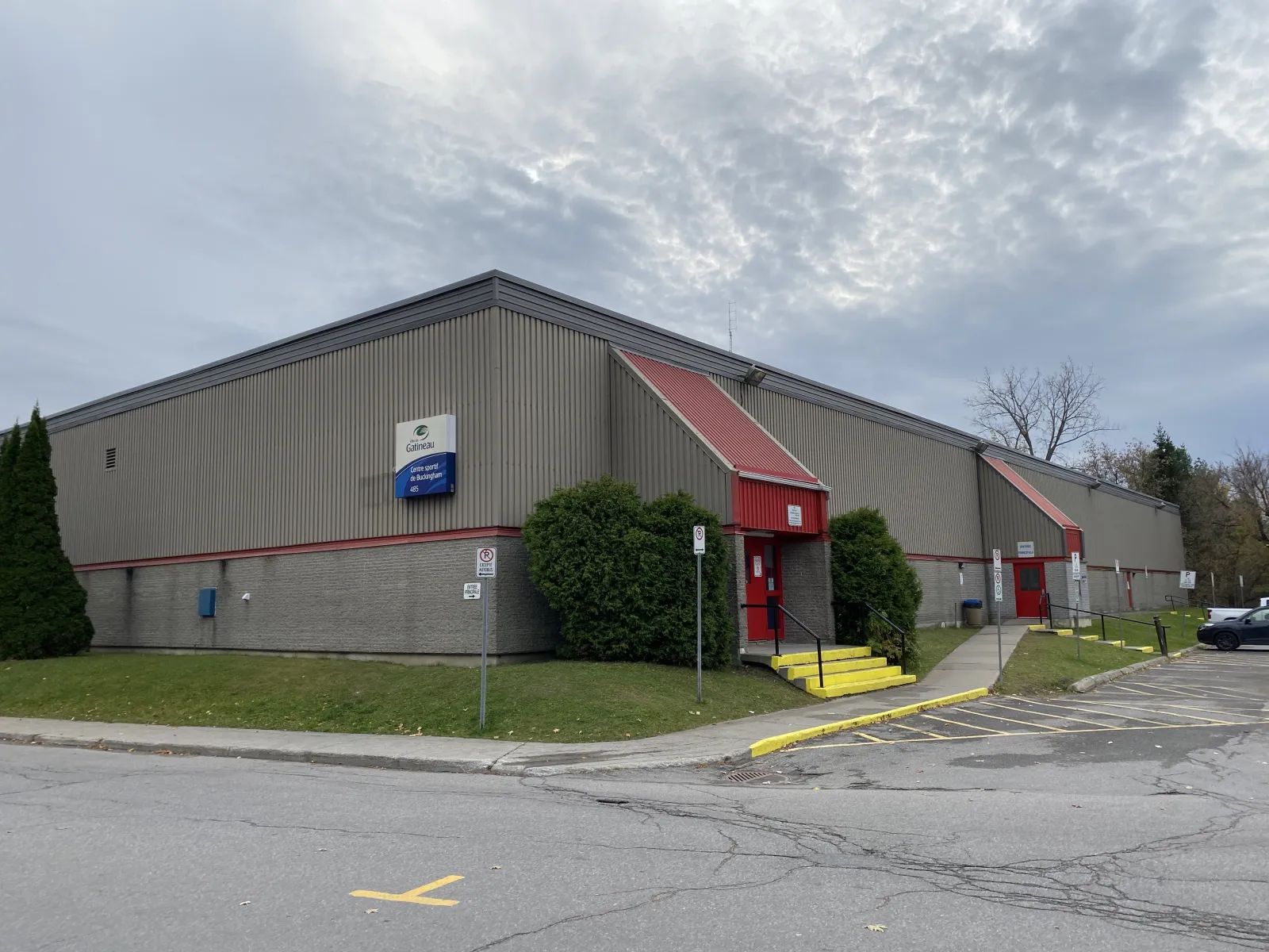 Le Centre sportif de Buckingham, un long aréna aux murs beiges dont les entrées latérales ont des toits rouges, avec une enseigne de la Ville de Gatineau portant le numéro 485. — Centre sportif de Buckingham