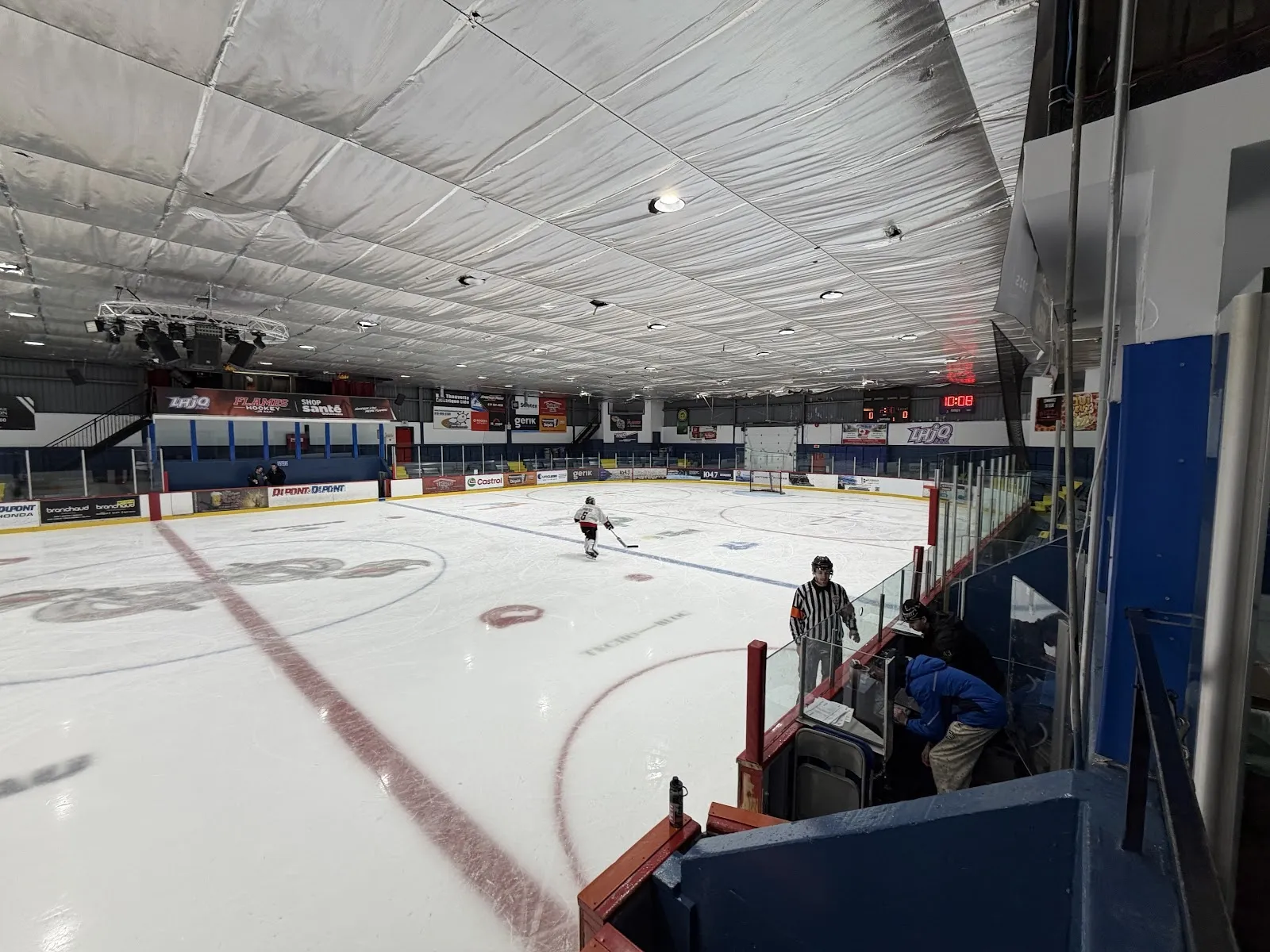 Une vue large le long de la glace avec un joueur seul au loin, un plafond gris nervuré au-dessus et une rampe rouge avec des bandes bleues du côté proche. — Centre sportif de Buckingham