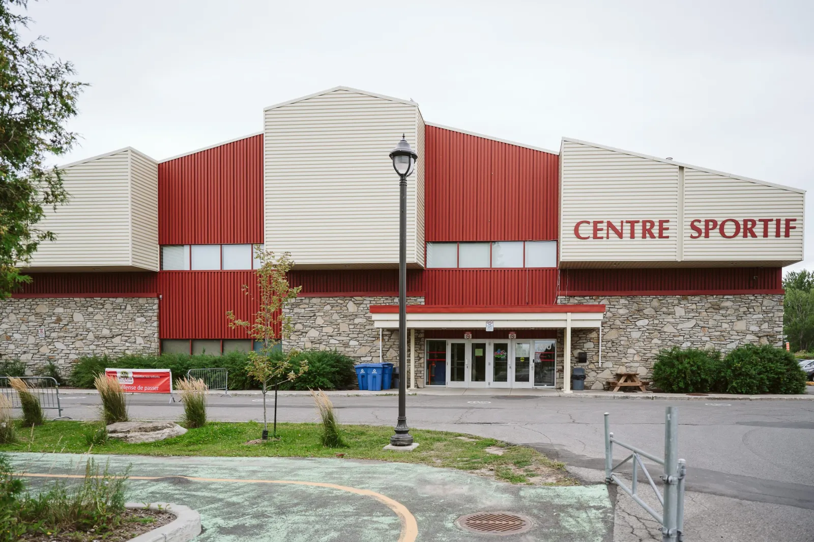 L’entrée principale du Centre sportif sous une marquise plate, avec une base de pierre, un revêtement métallique rouge et beige et le lettrage rouge CENTRE SPORTIF sur le haut du mur. — Centre sportif de Delson