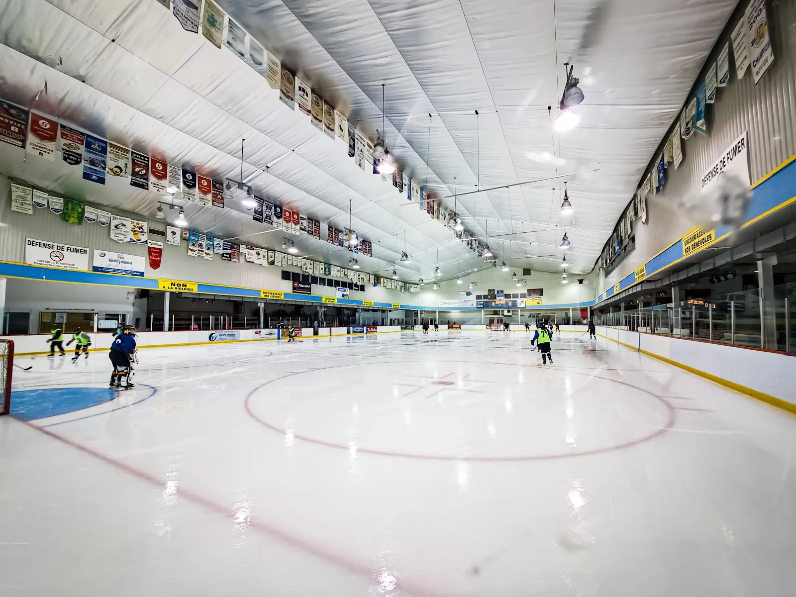 Toute la surface de glace vue d’un coin, au niveau de la glace, sous un plafond voûté blanc orné de bannières d’équipes, avec quelques jeunes joueurs de hockey qui patinent au loin. — Centre sportif de Delson