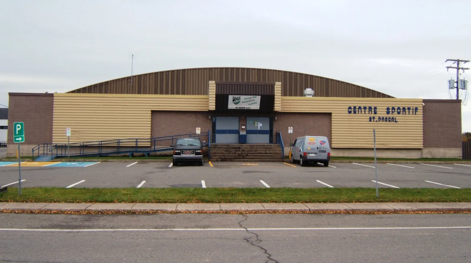 The front of the Centre sportif de Saint-Pascal across its parking lot, with the curved roof, the entrance stairs and the building name on the wall. — Centre sportif de Saint-Pascal
