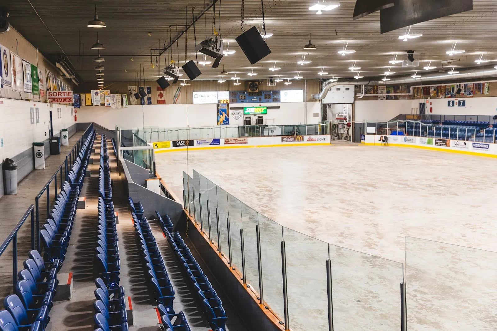 The ice sheet of the Centre sportif de Saint-Pascal seen from the stands, with blue seats along the glass and banners hanging above the far boards. — Centre sportif de Saint-Pascal