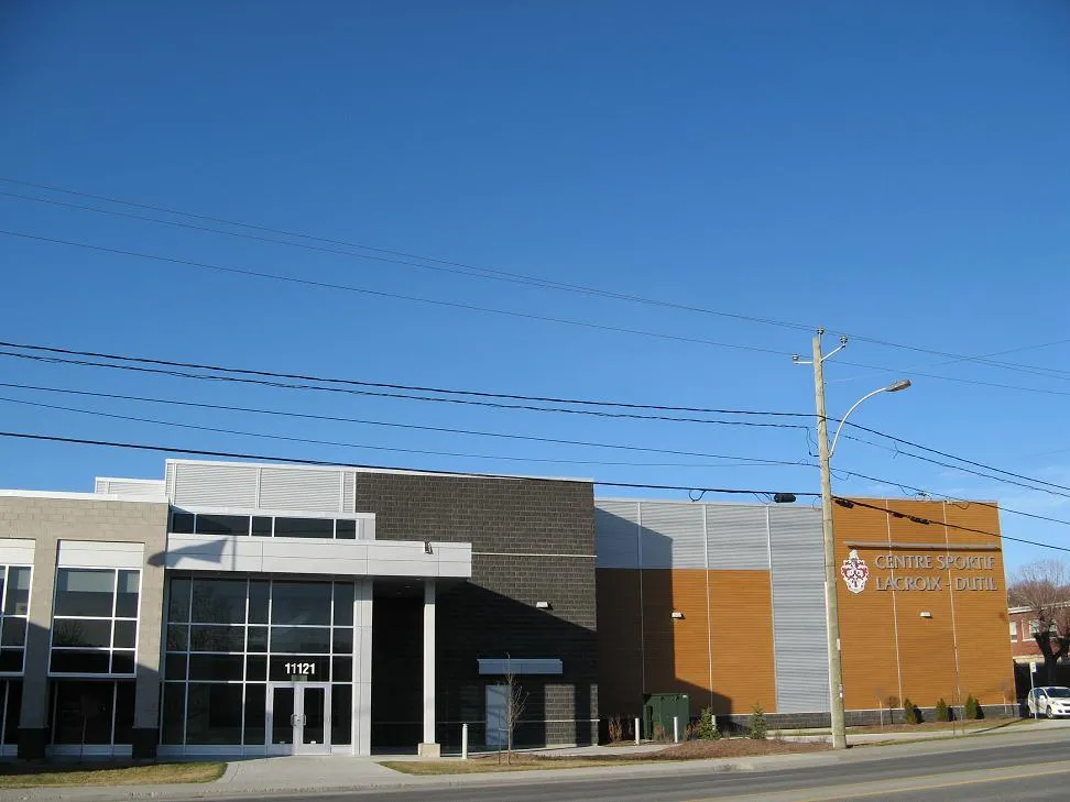 La façade du centre vue de la rue sous un ciel bleu : une entrée vitrée avec le numéro 11121 au-dessus de la porte, un revêtement gris et orangé de la couleur de la brique, et l’enseigne du Centre sportif Lacroix-Dutil à droite. — Centre sportif Lacroix-Dutil