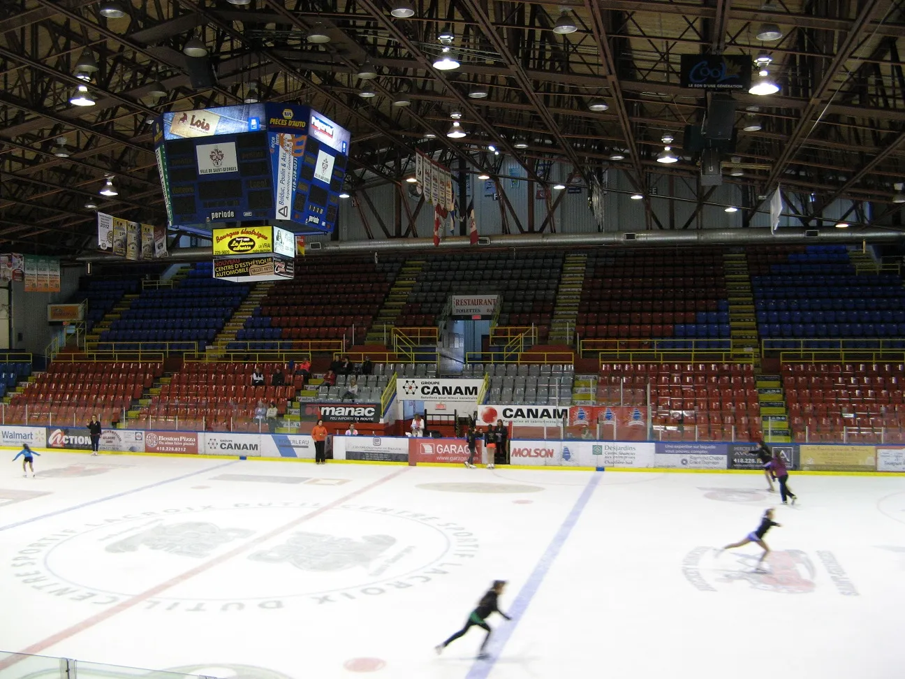 La glace Canam vue du niveau de la patinoire avec quelques patineurs, le logo du centre de la glace, les bandes Canam et Manac, les fermes d’acier du toit, le tableau d’affichage et les gradins rouges et bleus. — Centre sportif Lacroix-Dutil