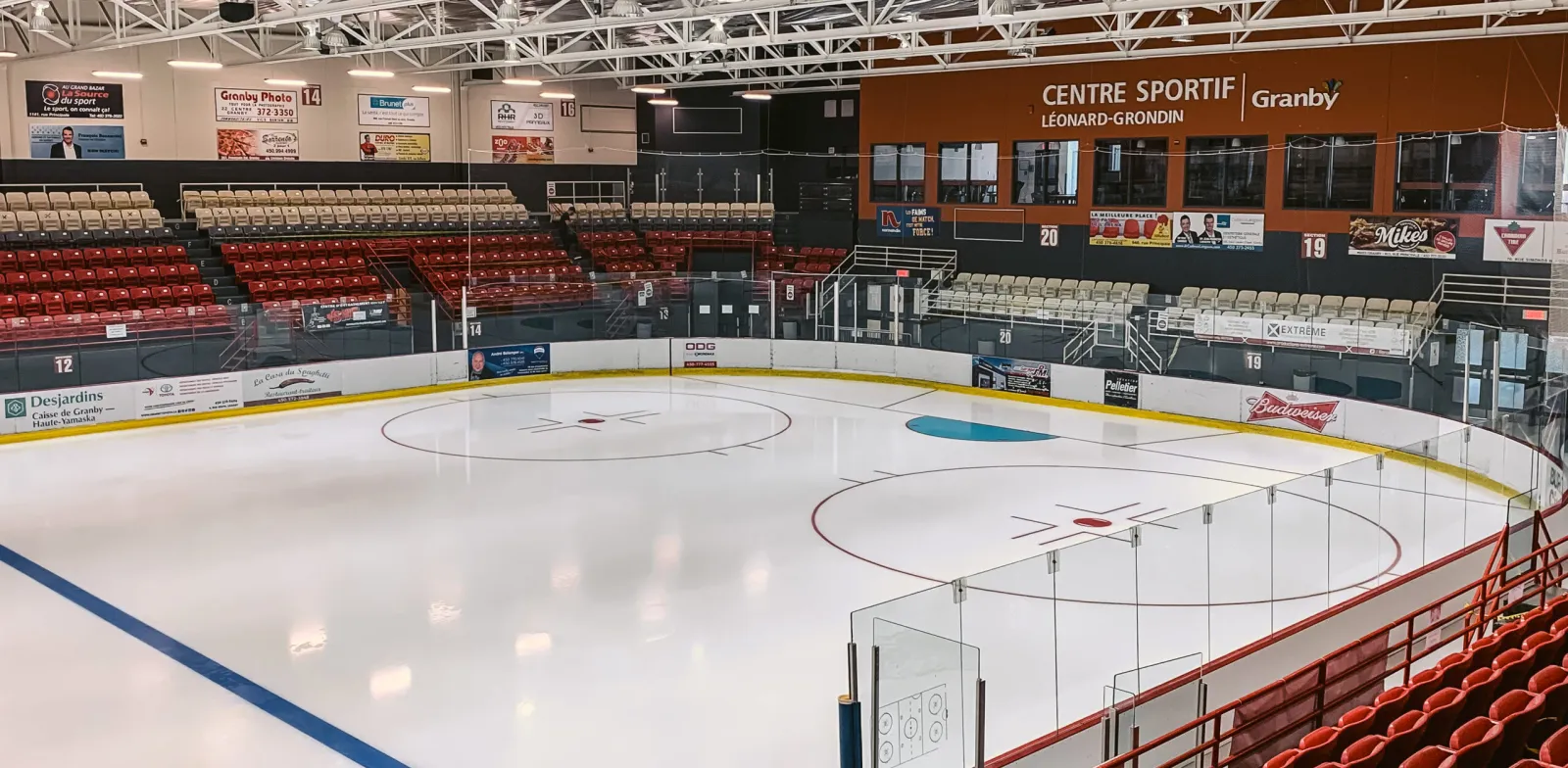 An empty sheet seen from the corner glass, with red seats along both sides and a Centre sportif Léonard-Grondin Granby sign above the far stands. — Centre sportif Léonard-Grondin