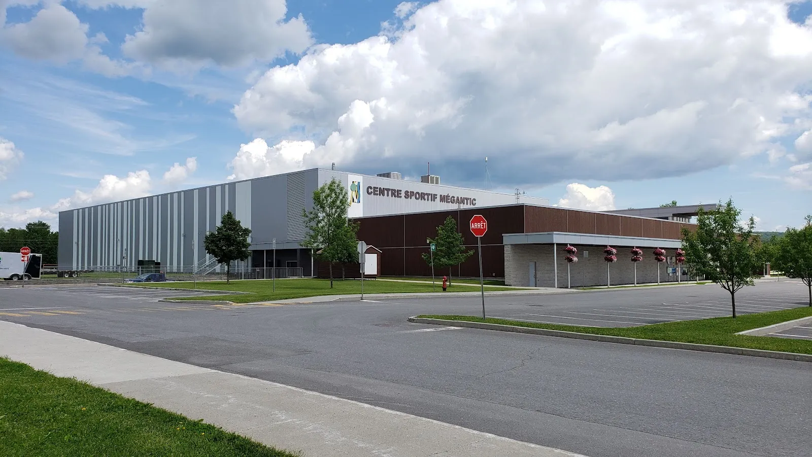 Le Centre sportif Mégantic vu de son stationnement : un haut volume gris abritant l’aréna, avec le nom du centre au-dessus de l’aile d’entrée en brique. — Centre sportif Mégantic