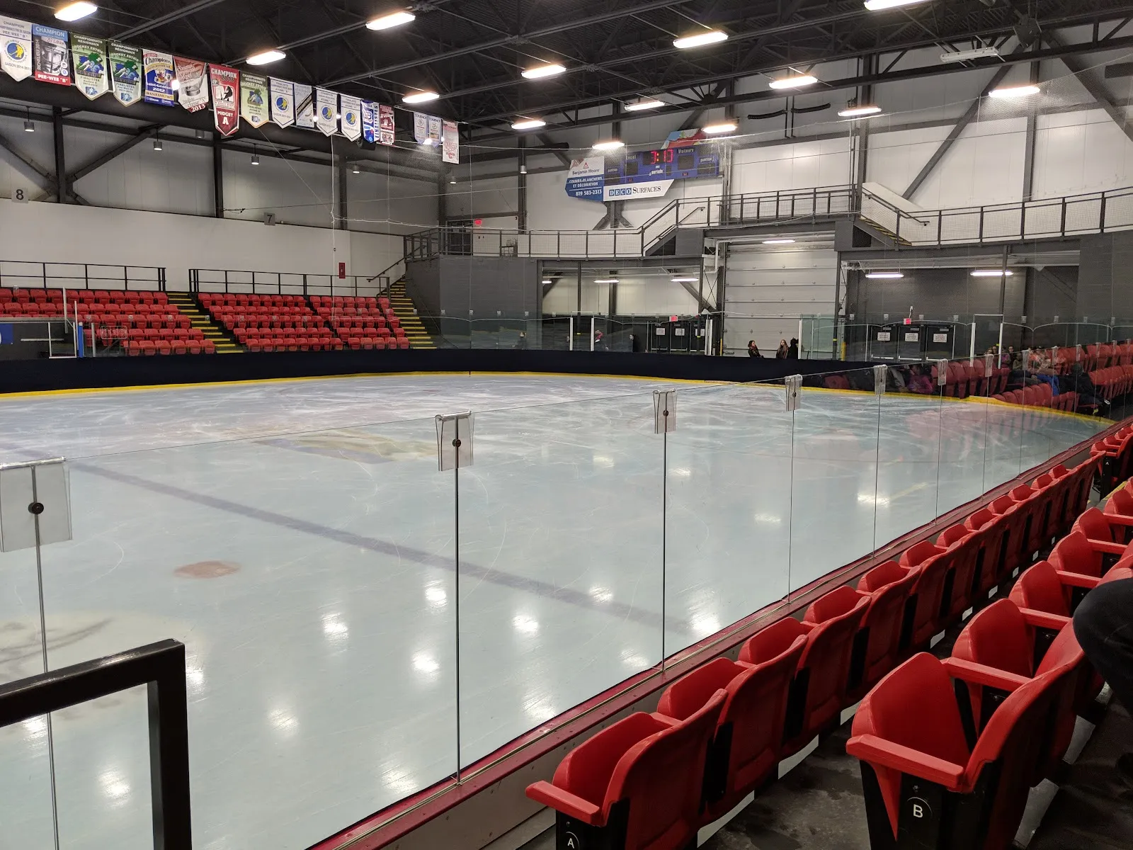 La patinoire vide vue au-delà des sièges rouges derrière la baie vitrée, avec des bannières d’équipes suspendues au toit d’acier blanc. — Centre sportif Mégantic