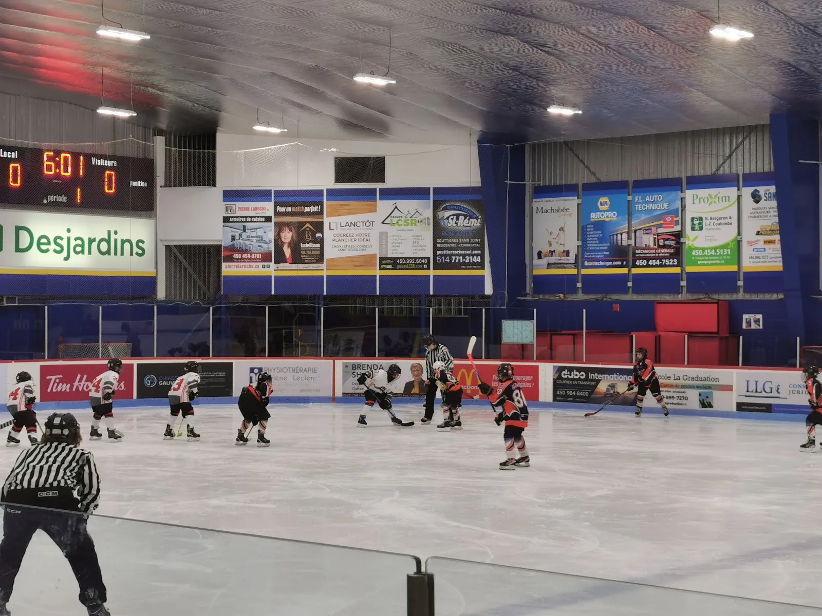 Un partido juvenil visto a través del vidrio, con mantas de patrocinadores en el muro azul del fondo y el marcador en la esquina. — Centre sportif régional Les Jardins du Québec