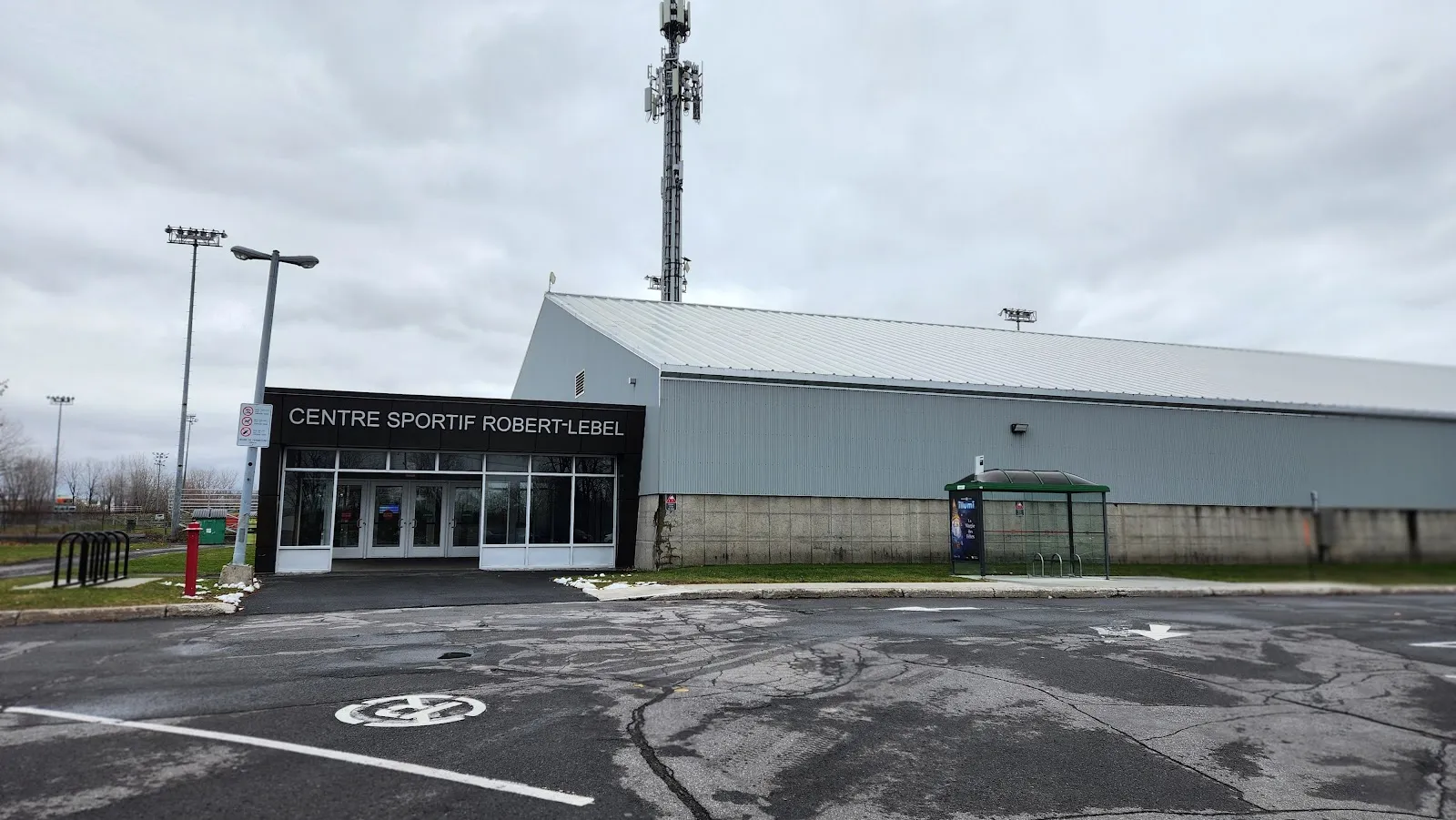 La entrada del Centre sportif Robert-Lebel bajo su rótulo, con el largo edificio gris revestido de metal del arena a su lado, una marquesina de autobús y el aparcamiento bajo un cielo nublado. — Centre sportif Robert-Lebel